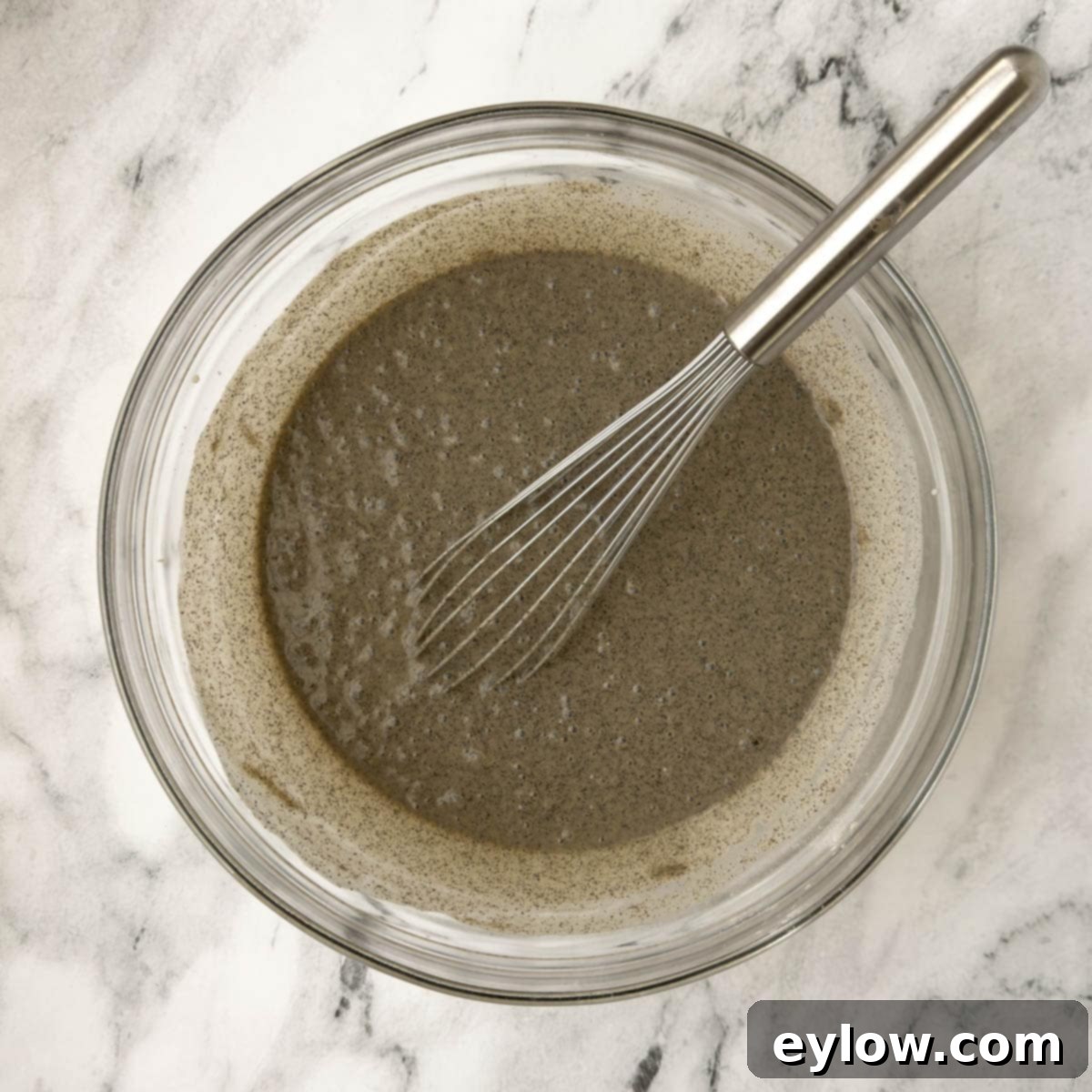 Rich, dark buckwheat pancake batter is thoroughly mixed in a glass bowl using a silver whisk, showcasing its smooth, consistent texture.