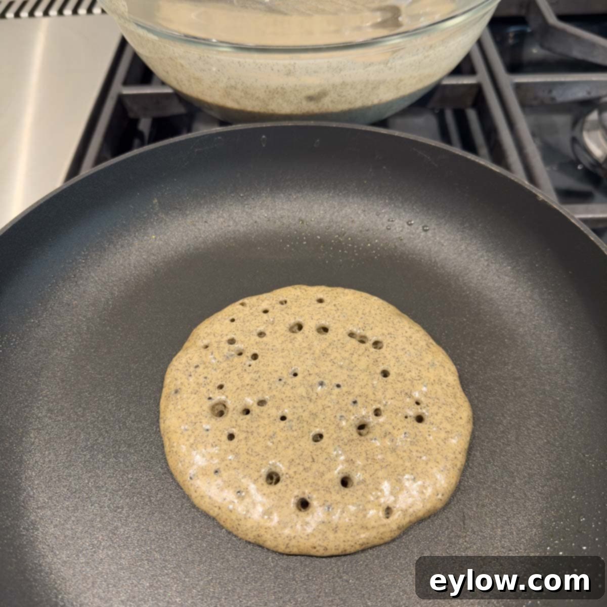 A single buckwheat pancake sizzling in a non-stick pan, showing small bubbles forming on its surface, indicating it's nearly ready to be flipped.