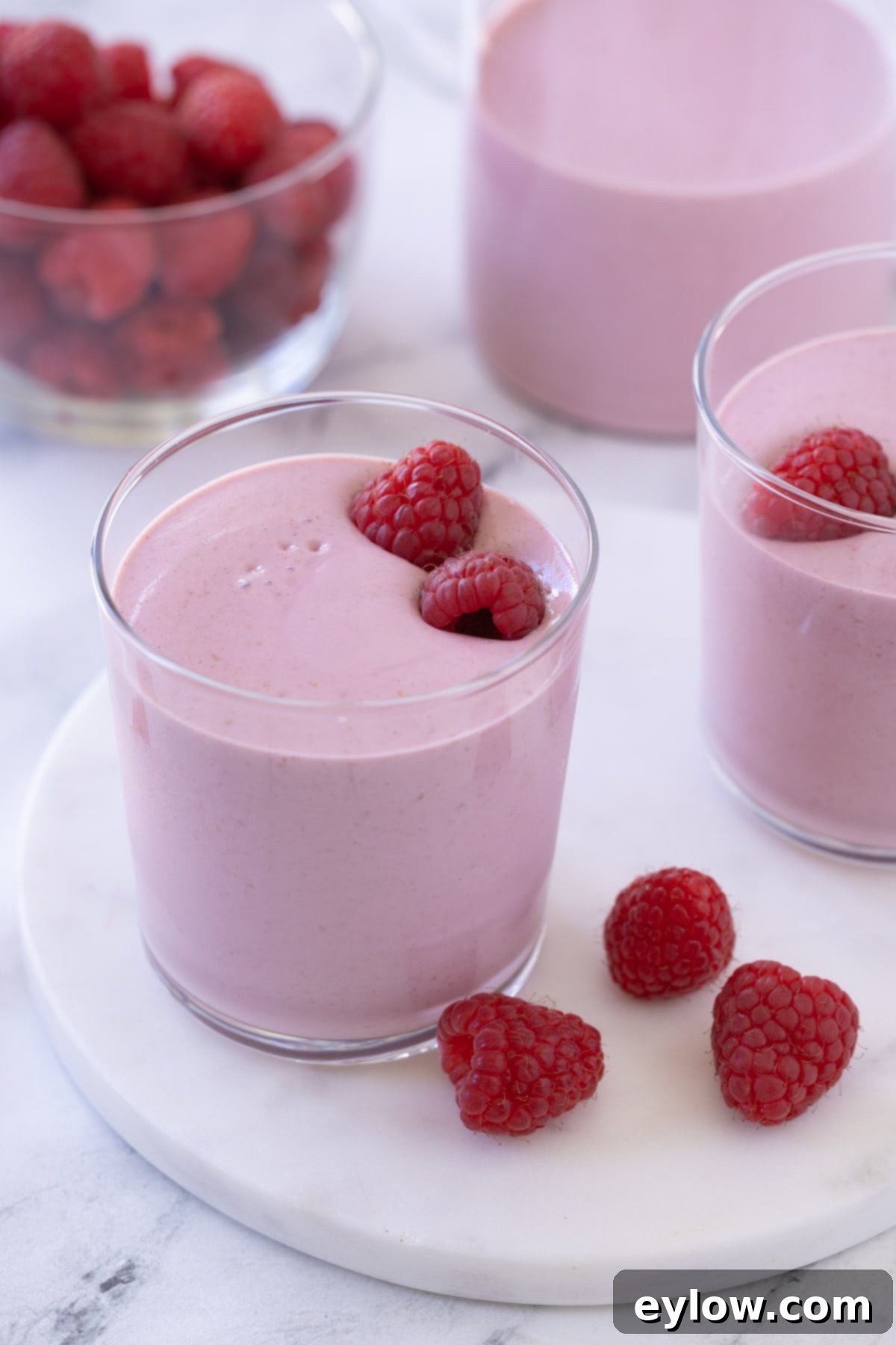 A beautiful pink raspberry smoothie in a small glass with a bowl of raspberries.