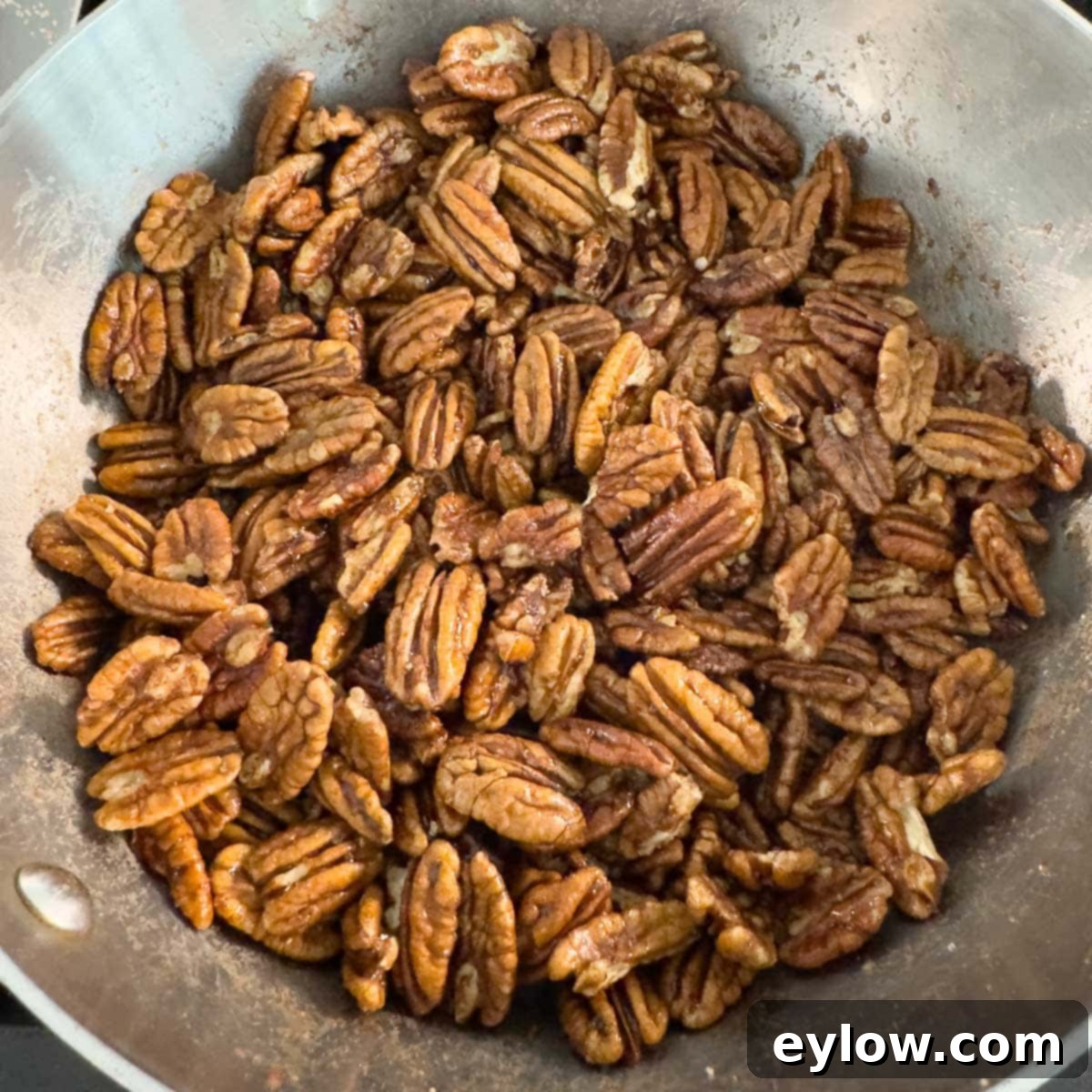 Whole pecans in a silver pot on the stovetop, being stirred to coat evenly with the maple glaze.