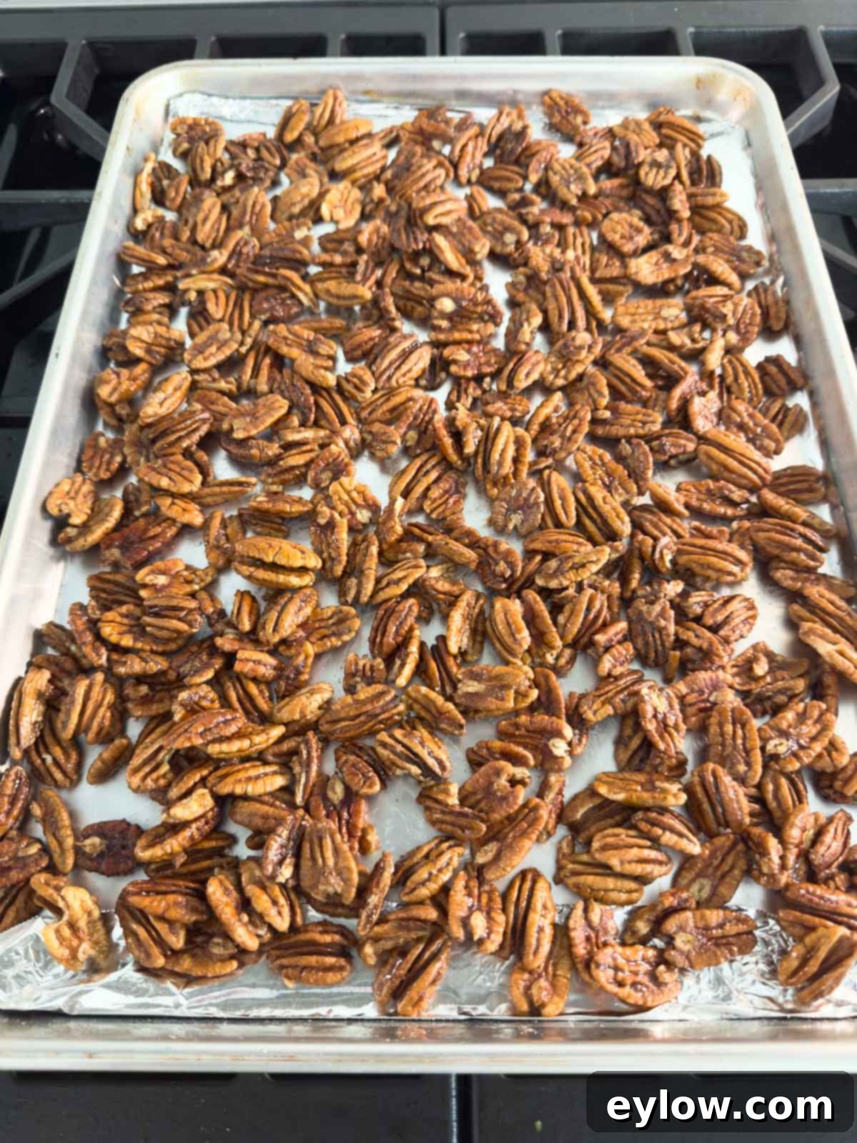 Whole pecans coated in maple syrup, butter, and brown sugar spread in a single layer on a rimmed baking sheet with foil.