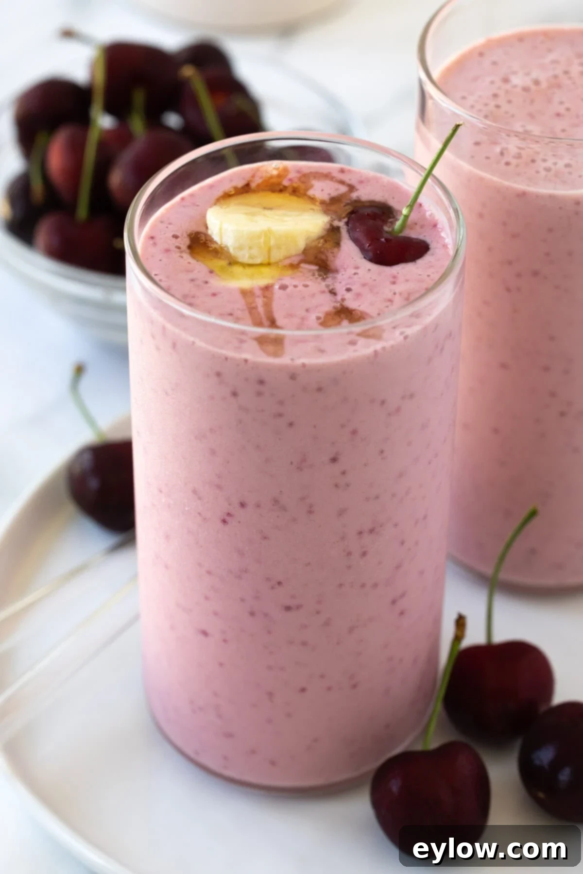 Easy 5-Minute Cherry Smoothie 2 Two glasses of bright pink cherry smoothie, garnished with fresh dark cherries and a banana slice, with a bowl of dark cherries in the background.