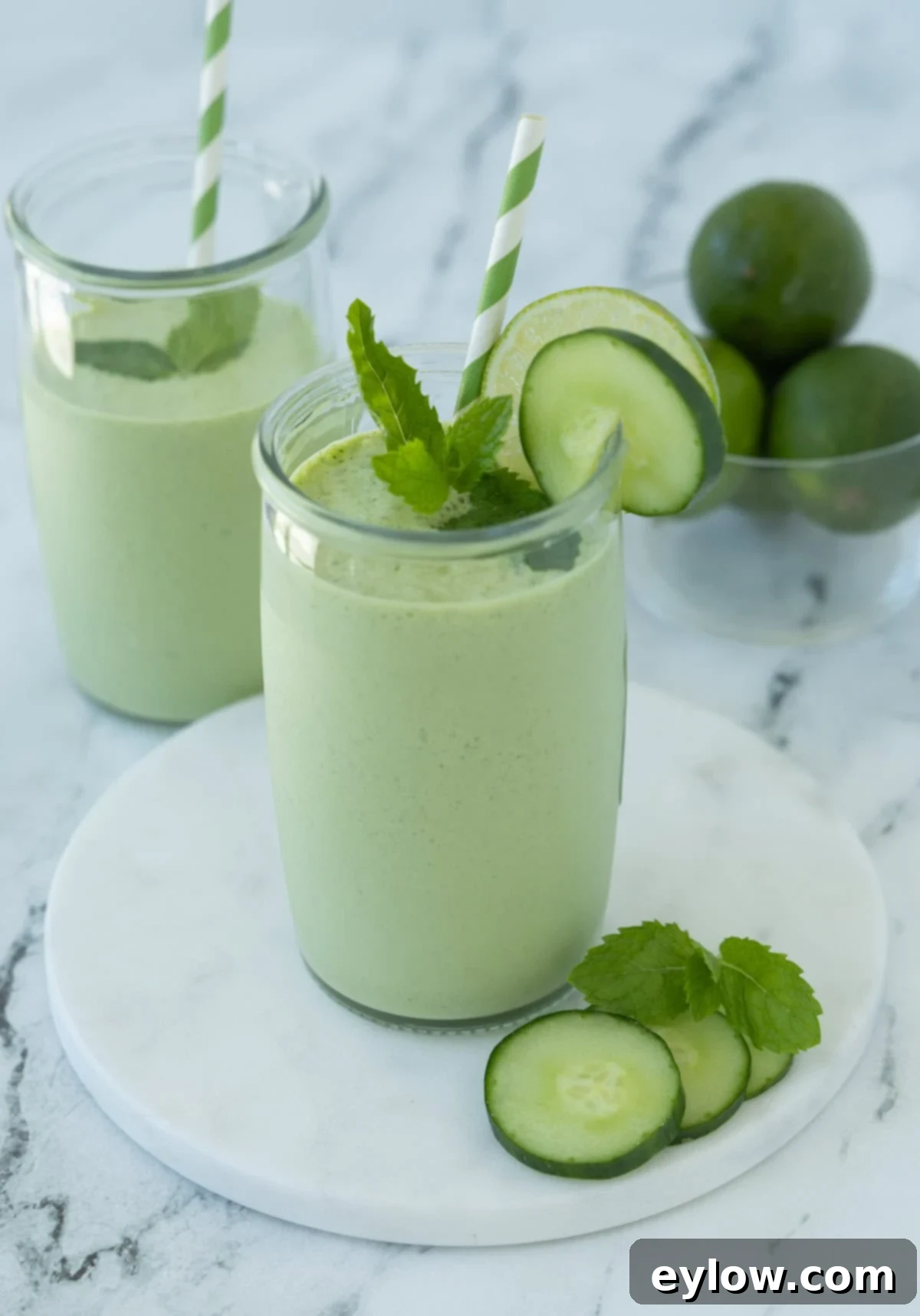 Minty green cucumber smoothie in a glass with mint leaves, cucumber slices, lime slice, and green striped straw.