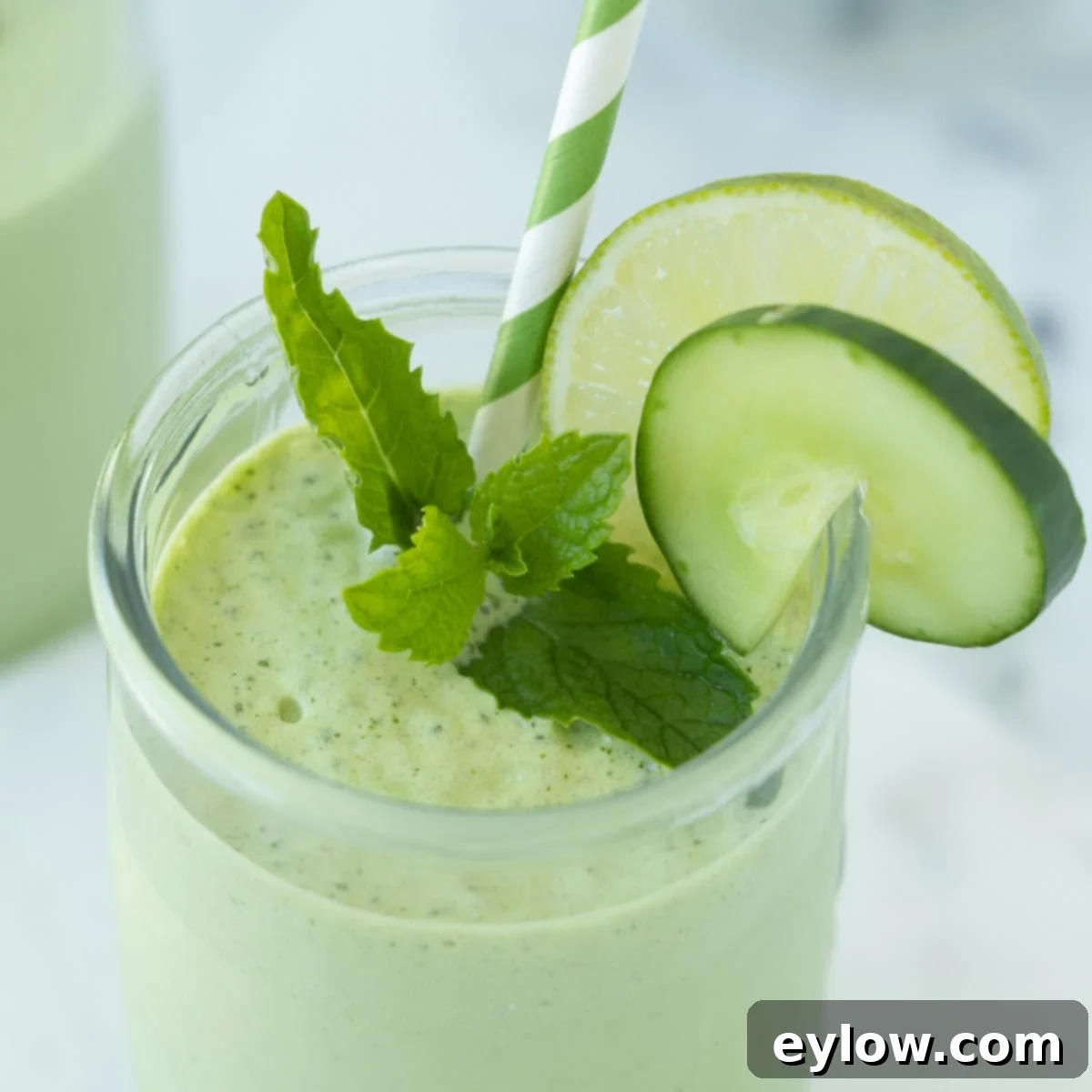 Creamy green smoothie with cucumber, mint, lime, and yogurt in a glass with a green striped straw, garnished with fresh mint.