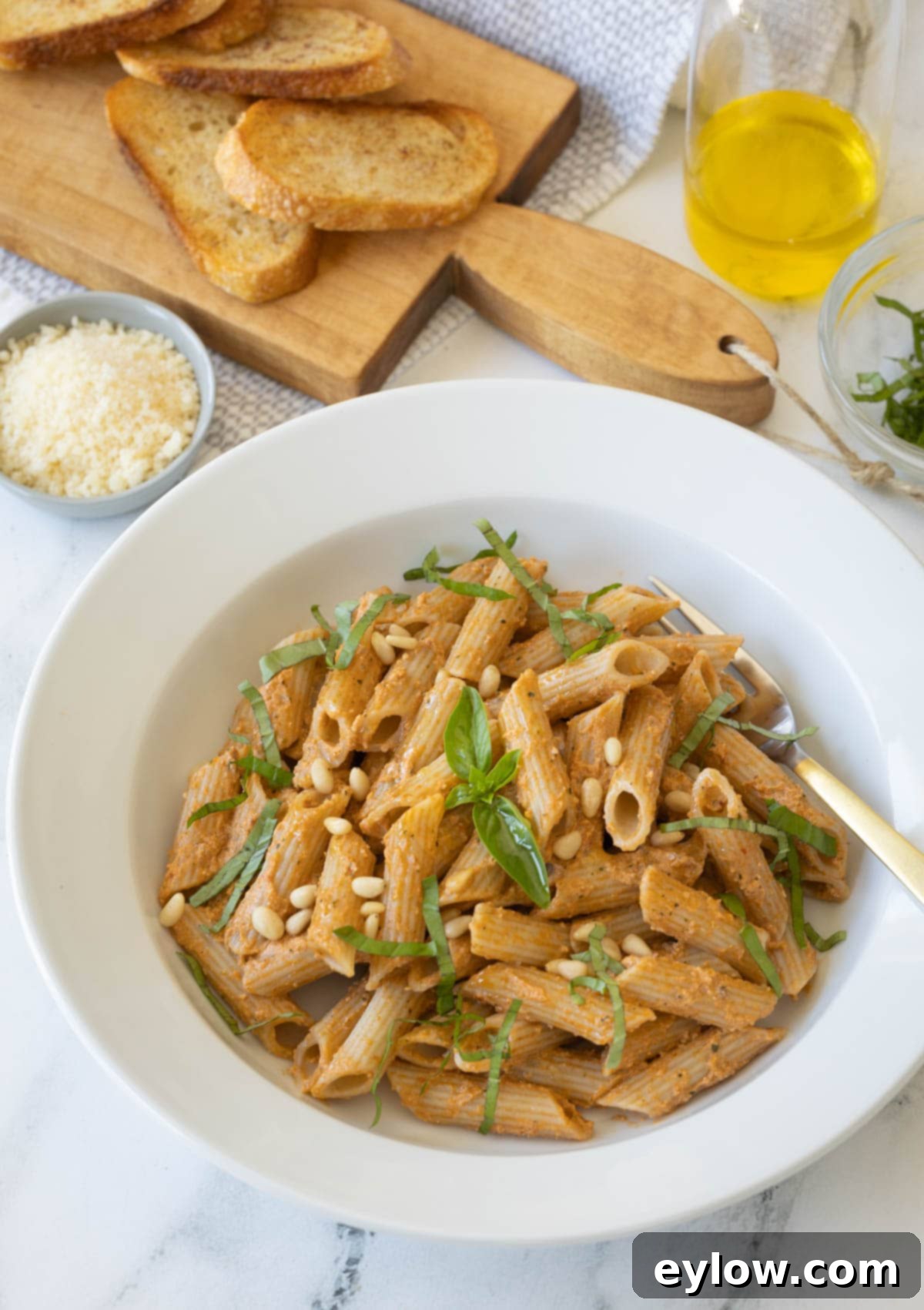 Velvety Sun-Dried Tomato Pesto Pasta 2 A white bowl of creamy sun-dried tomato pesto pasta with fresh basil leaves, pine nuts, toasted bread, and grated parmesan.