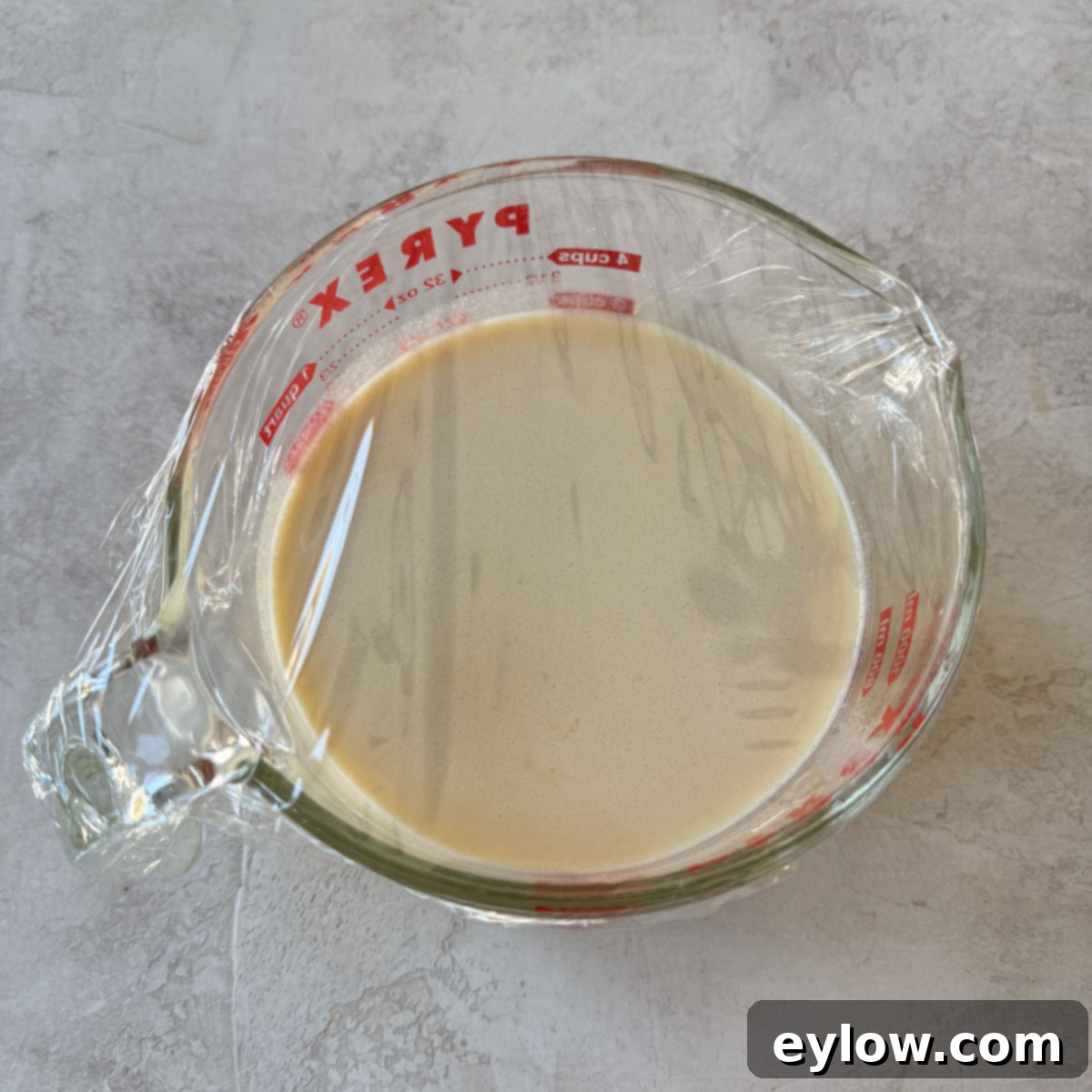 A Pyrex glass measuring container filled with light yellow crepe batter, covered with plastic wrap and ready for resting.
