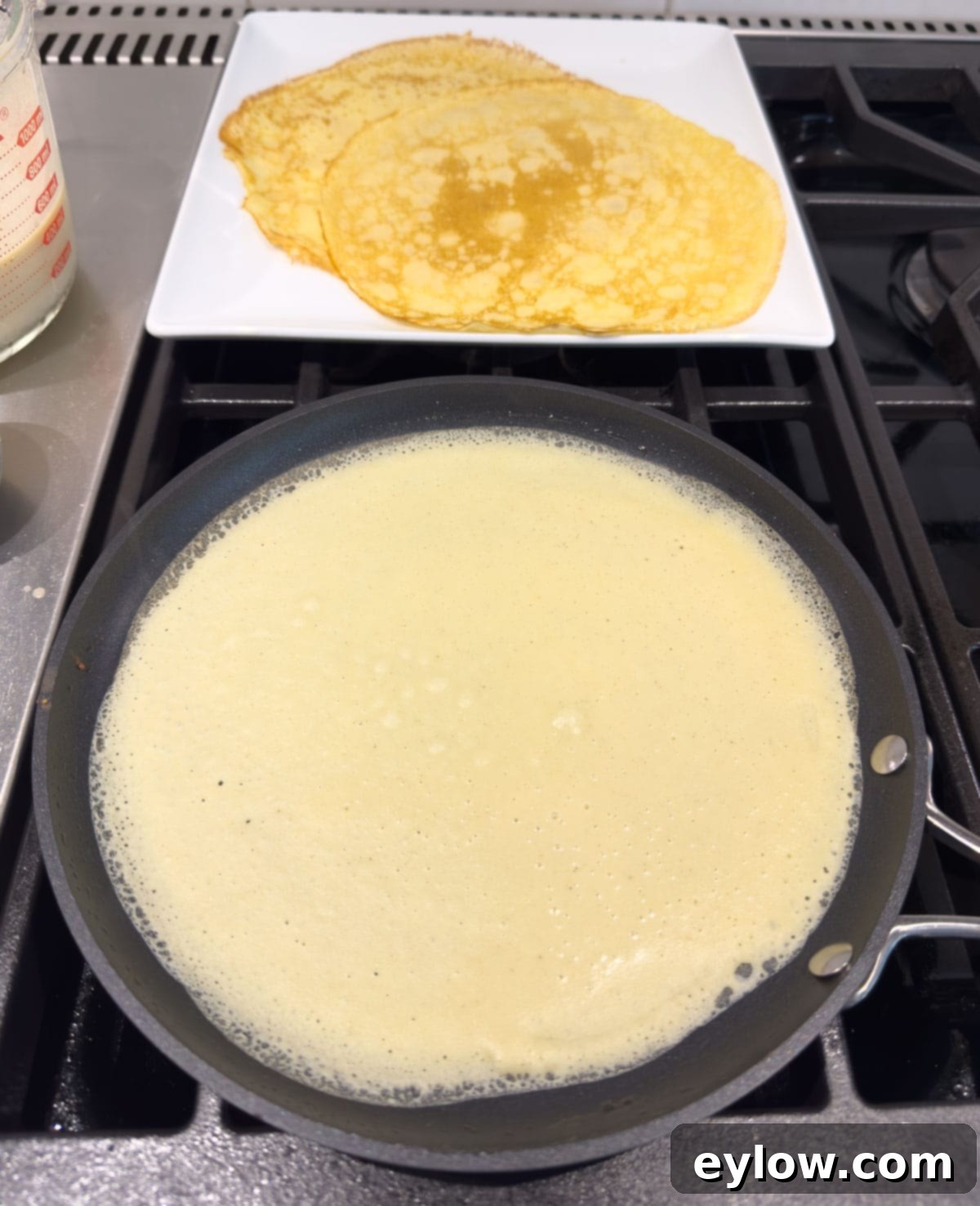 Crepe batter being swirled in a crepe pan, with a stack of finished crepes visible on a nearby plate.