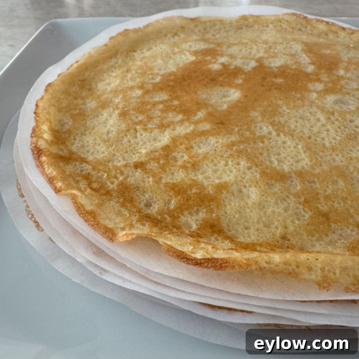 A neatly stacked pile of golden crepes, separated by rounds of parchment paper, on a white plate, indicating readiness for storage or serving.
