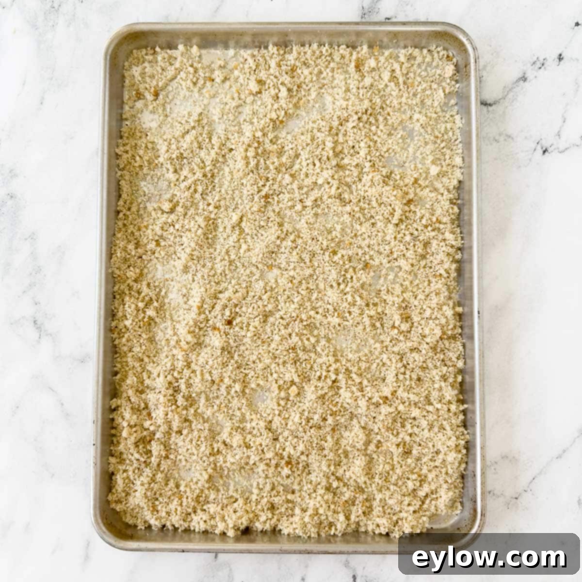 Raw gluten-free bread crumbs scattered evenly on a rimmed baking sheet, prepared for baking and drying.
