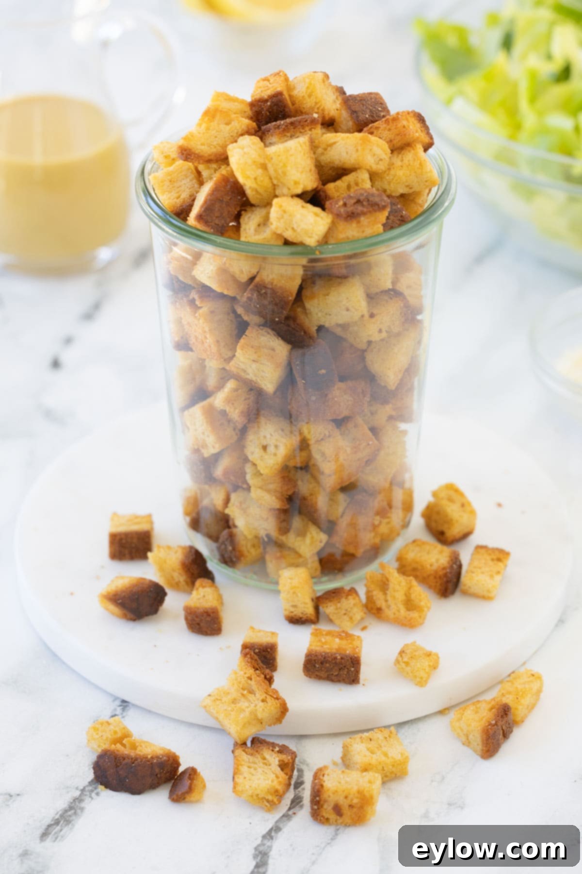 A tall glass jar of golden brown homemade croutons with some on the table.