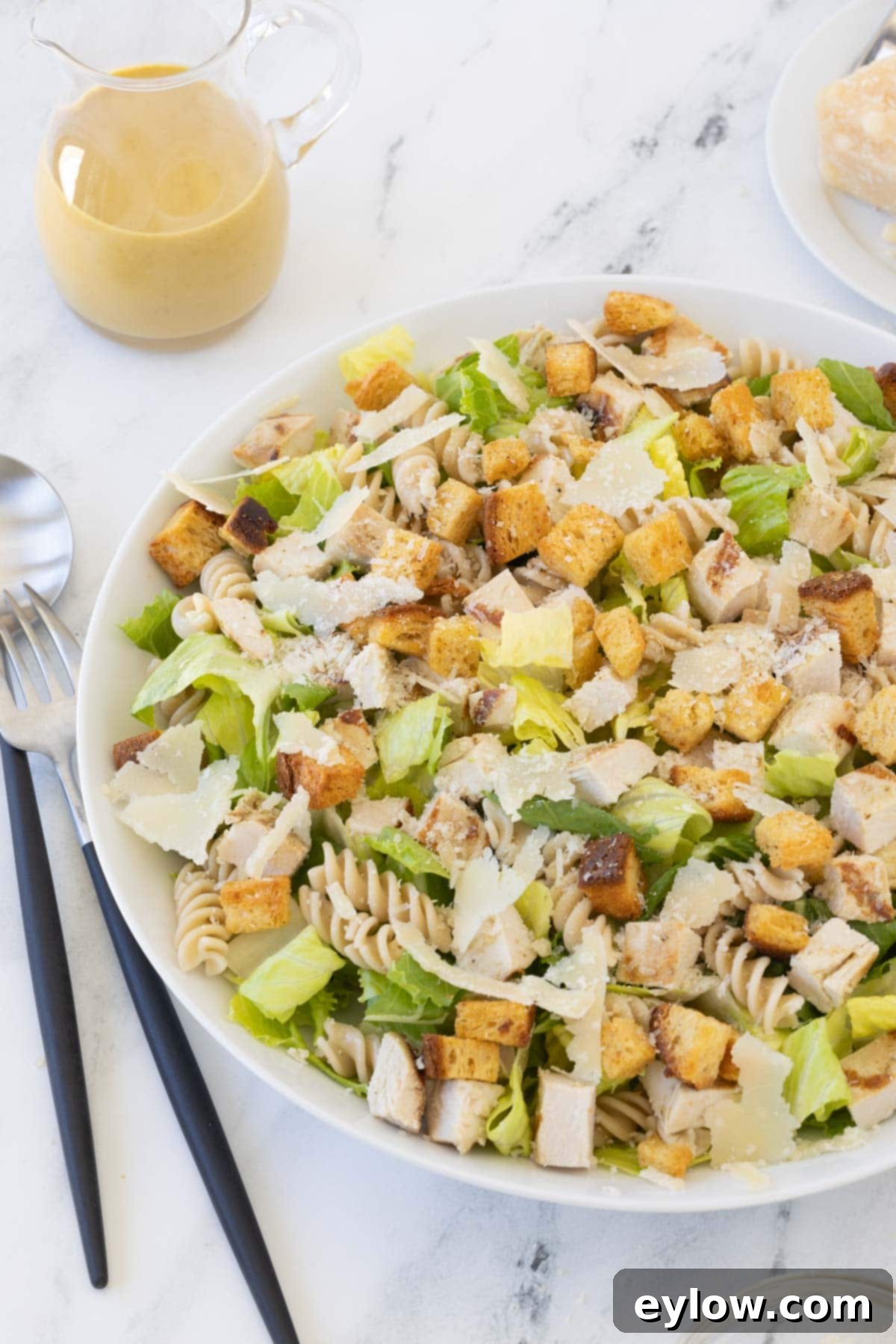 Emperor's Chicken Pasta Salad 2 A large shallow bowl of caesar pasta salad with romaine, curly pasta, croutons, chicken and dressing in a small pitcher.