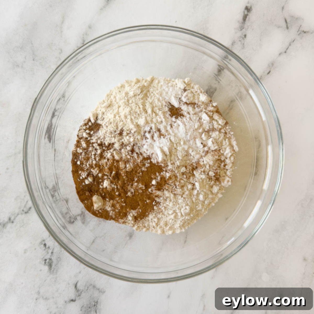 Dry ingredients for pumpkin bread, including gluten-free flour and a blend of warm spices, expertly sifted into a clear glass bowl, showcasing their readiness for mixing.