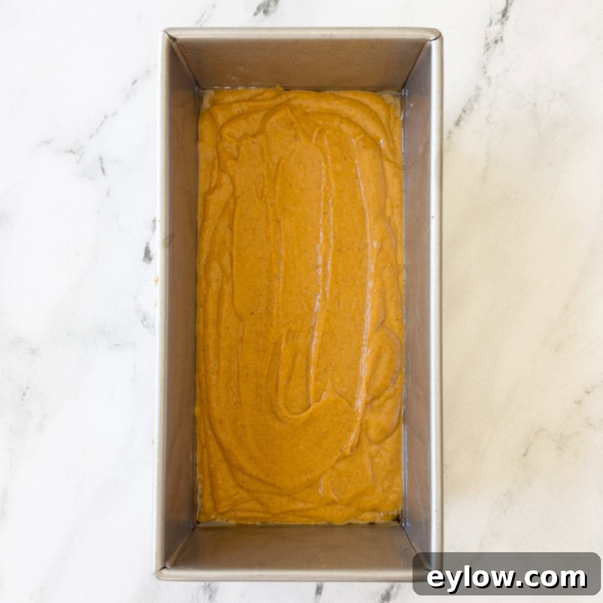 The rich, smooth pumpkin bread batter meticulously poured into a standard silver metal baking pan, lightly sprayed with cooking spray, perfectly prepared and ready for the oven.