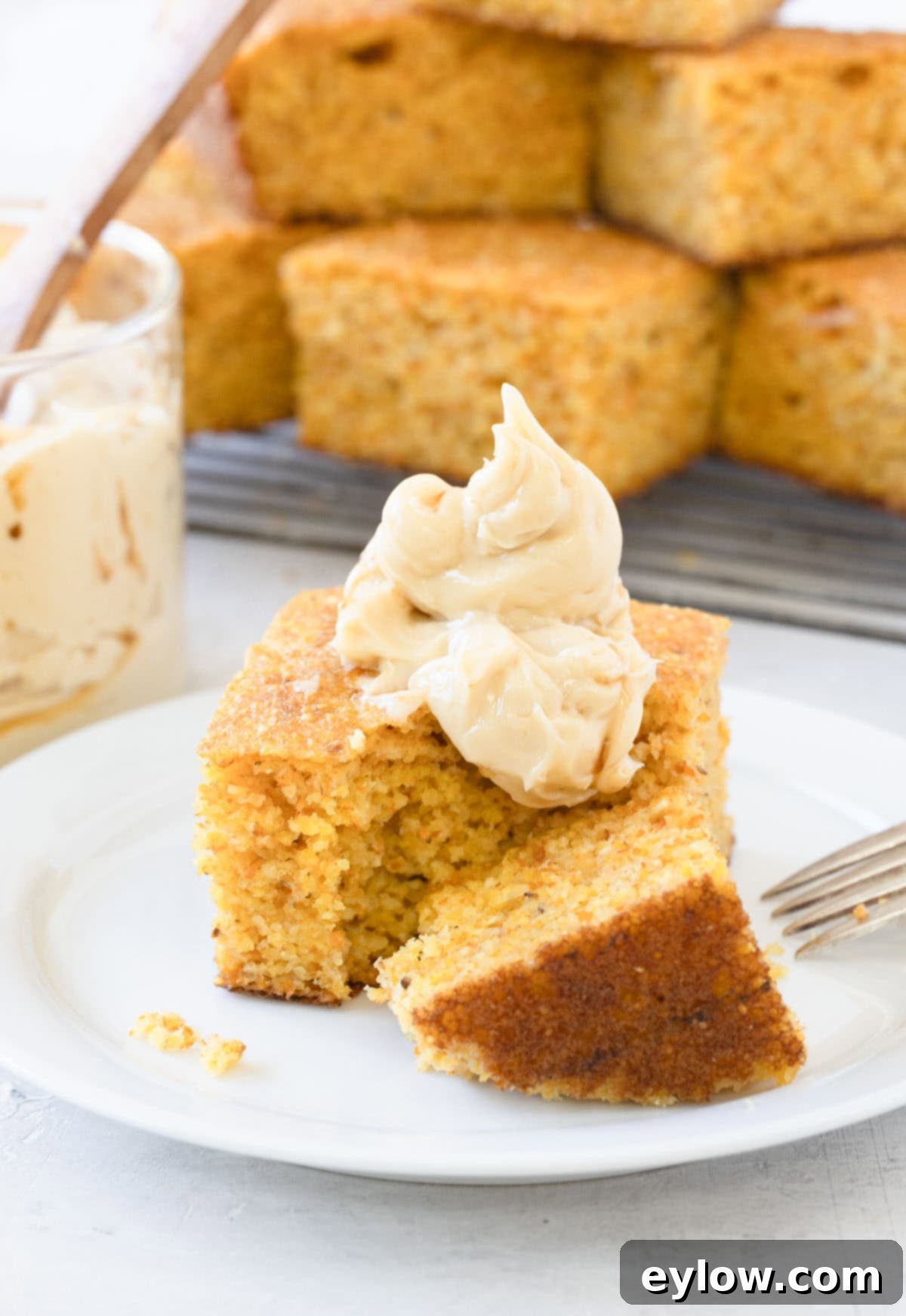 A slice of golden cornbread topped with a generous dollop of maple butter, melting slightly, on a white plate with a fork.