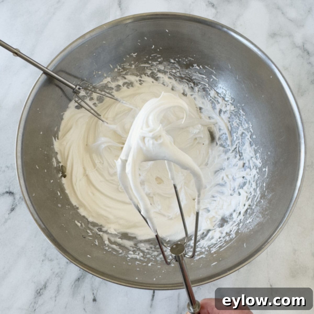 A hand mixer's beater covered in perfectly whipped, stiff egg white meringue over a steel mixing bowl, demonstrating the correct consistency.