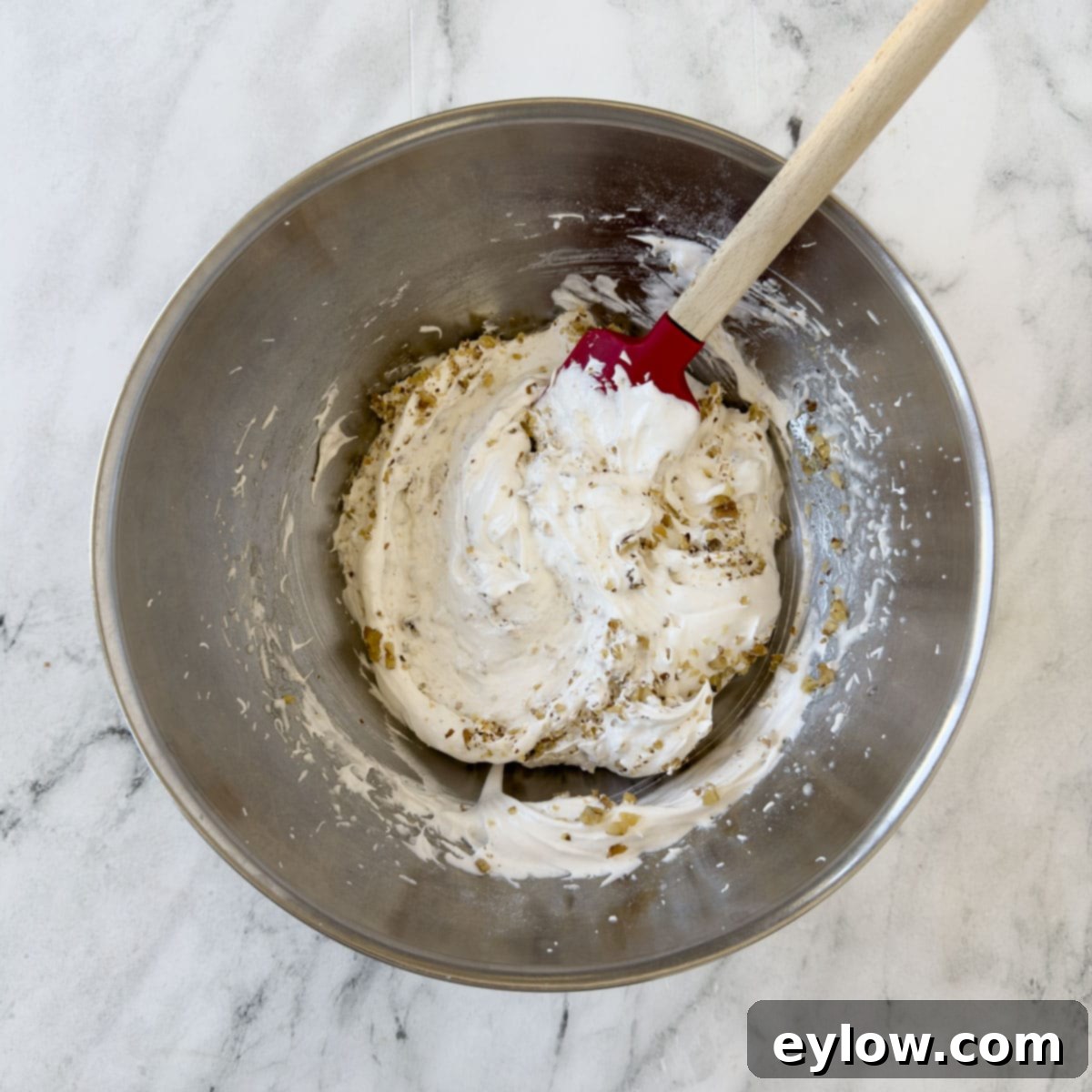 A rubber spatula gently folding finely chopped nuts into the stiffly whipped egg white meringue batter in a bowl.