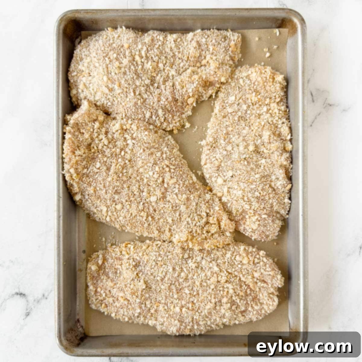 Four chicken cutlets coated in seasoned panko crumbs on a rimmed baking sheet.