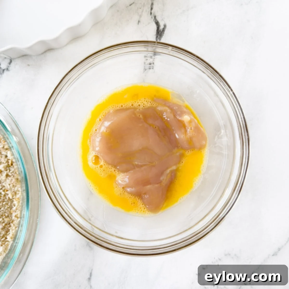 Whisked egg in a glass bowl with a chicken cutlet being coated.