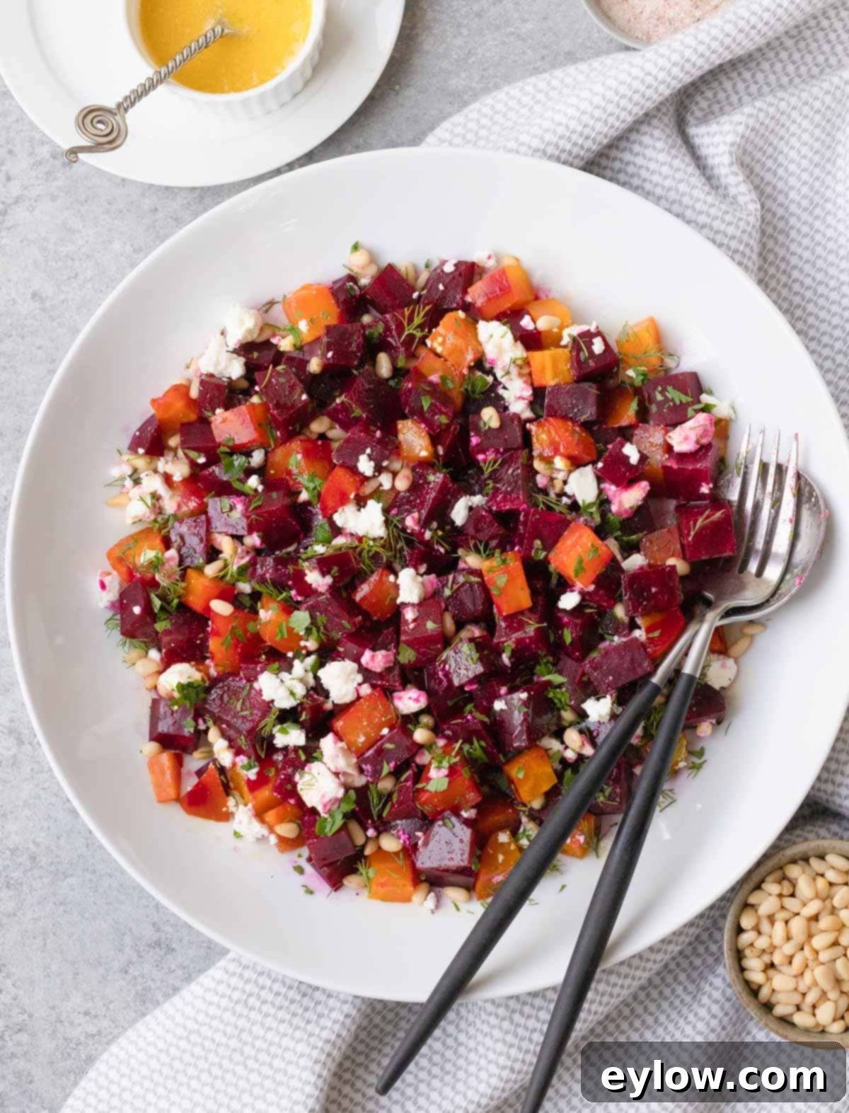 A white bowl of jewel-colored beet salad, generously topped with crumbled feta cheese, golden pine nuts, and fresh green herbs, ready to be served.