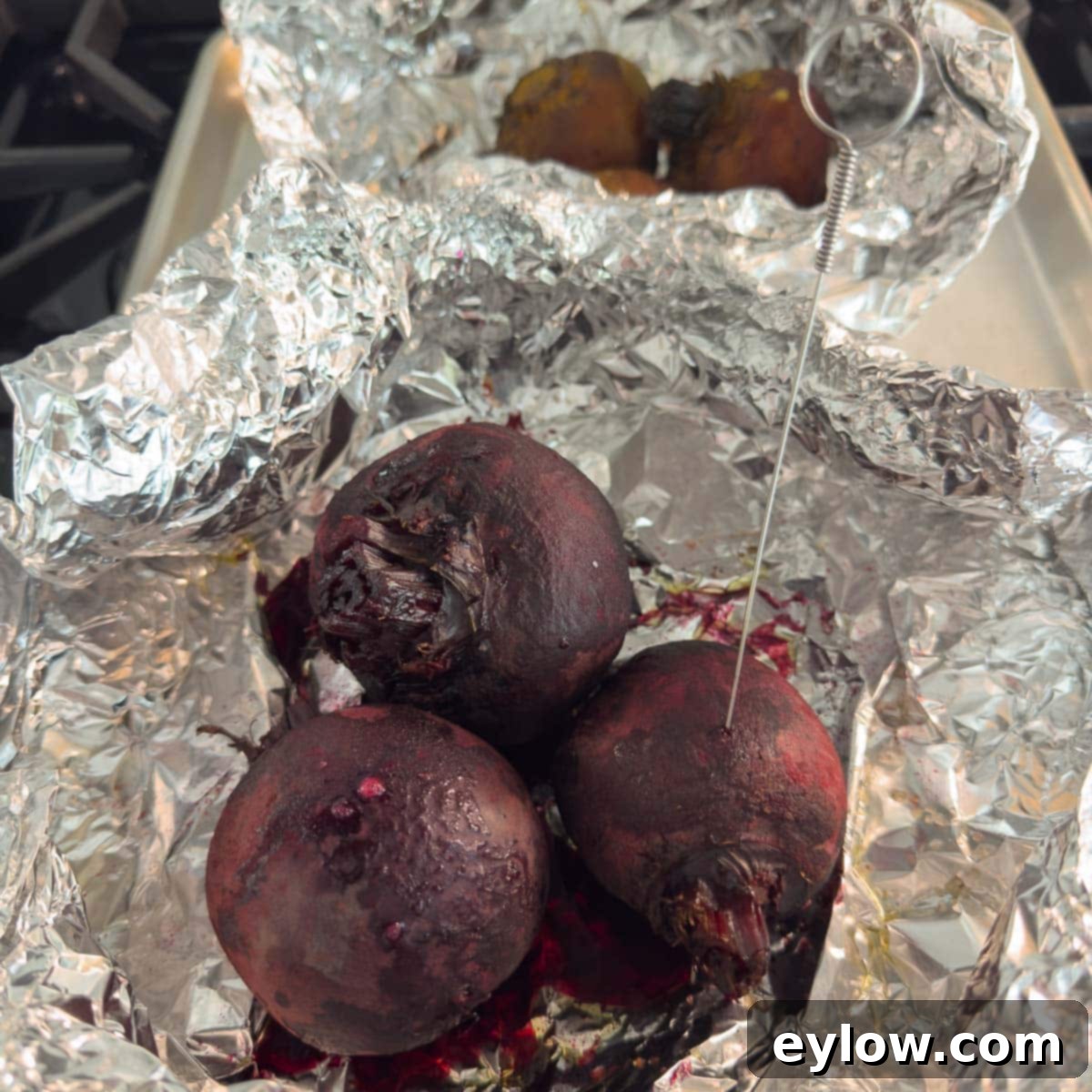 Roasted beets, still in their aluminum foil packets, with a thin metal skewer piercing one beet to show it's perfectly tender and done.