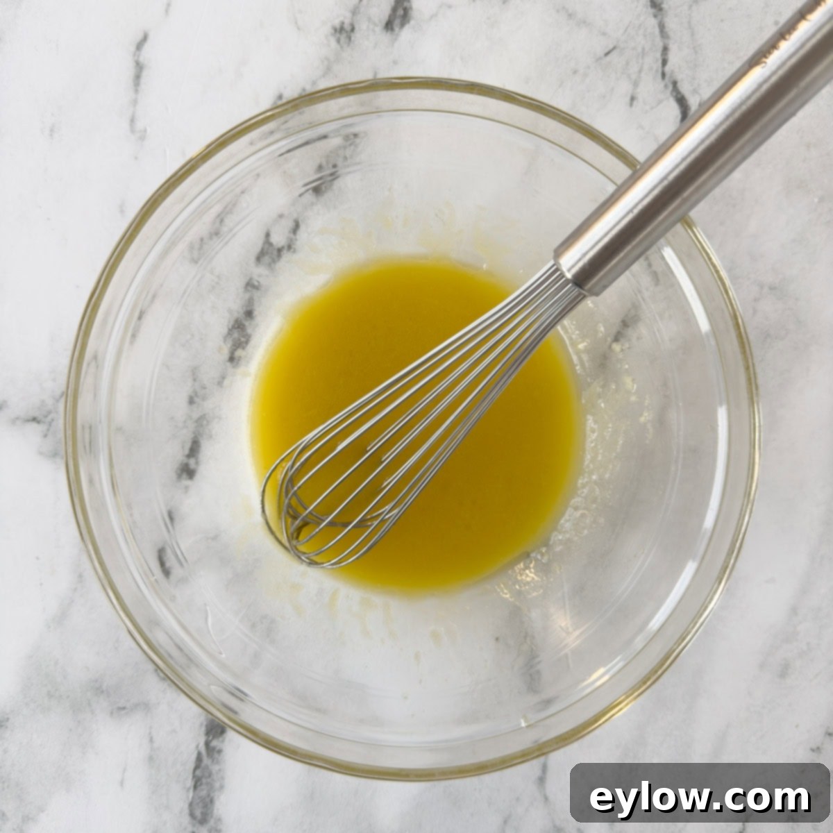 A homemade vinaigrette salad dressing being whisked vigorously in a clear glass bowl with a silver whisk, showing the emulsification process.
