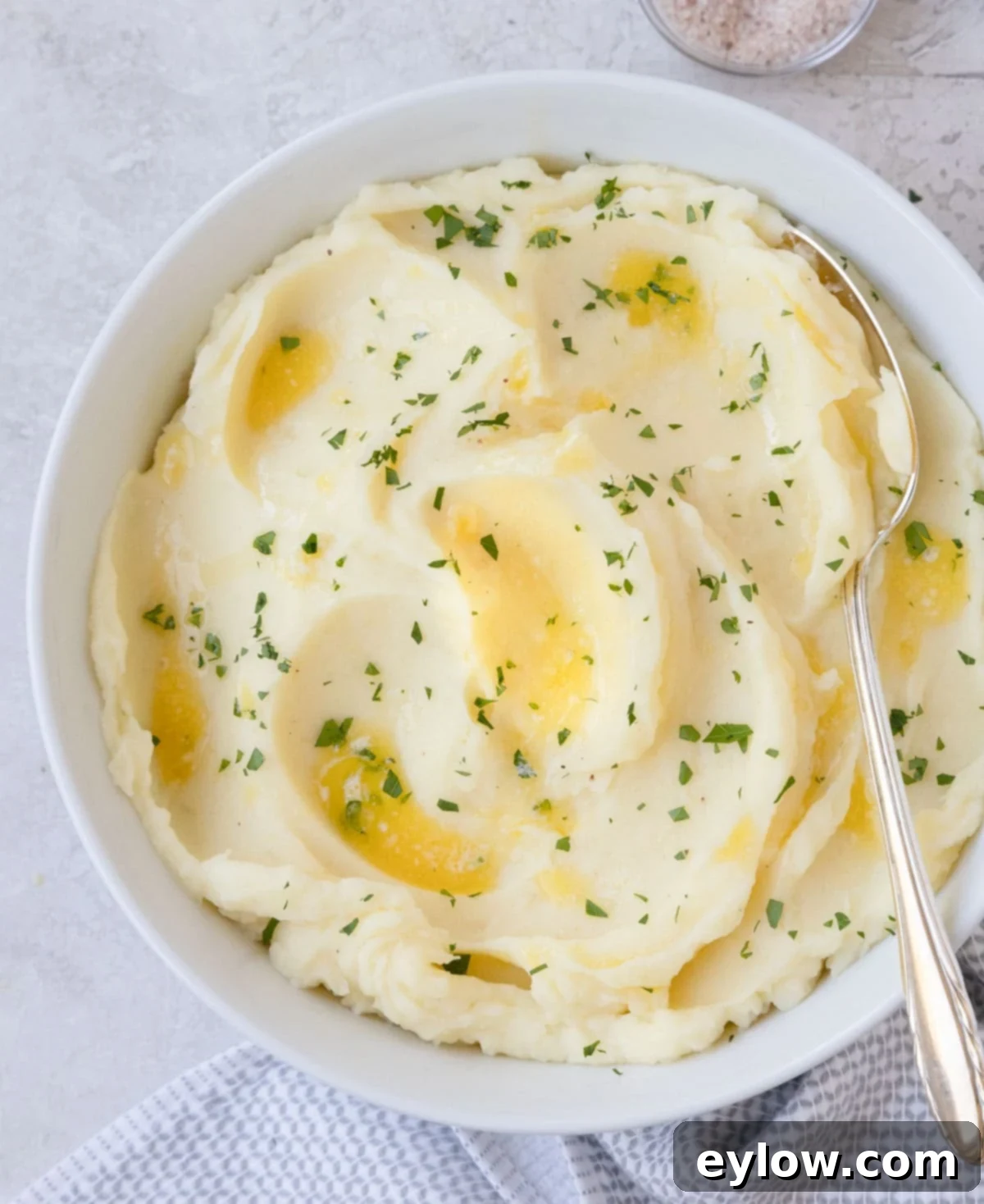 A round white bowl full of creamy mashed potatoes drizzled with melted butter and fresh chopped parsley.