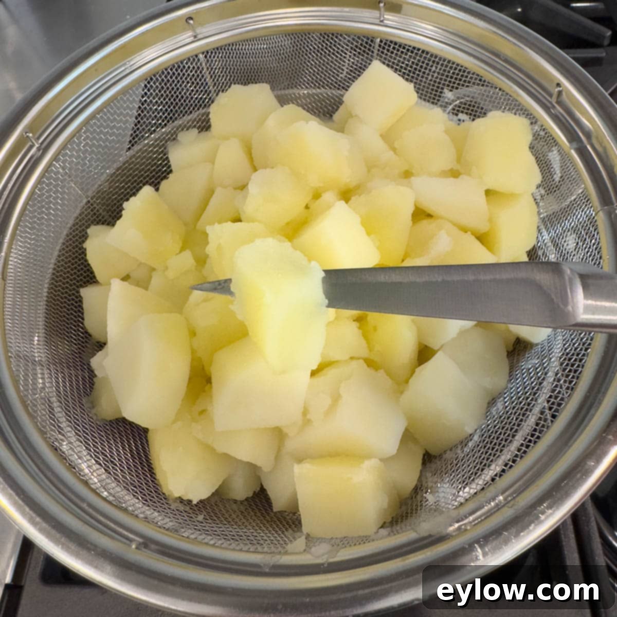 Cooked potatoes in a colander pierced with a paring knife showing doneness. 