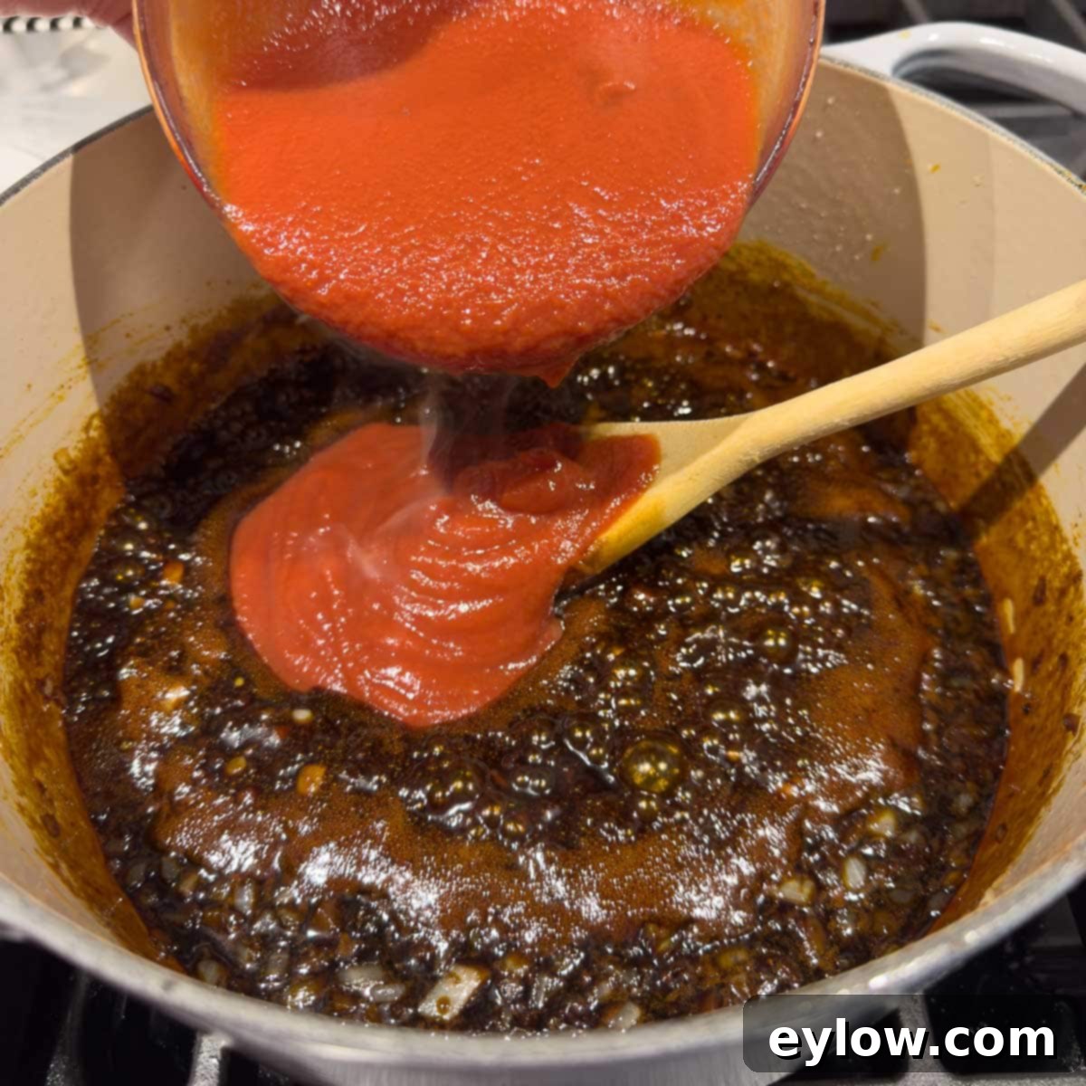 Pouring bright red ketchup into a pot with dark brown molasses and tender navy beans, mixing into the sauce.