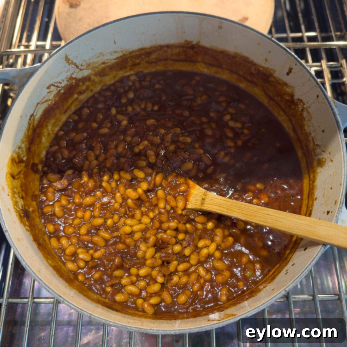 Tender navy beans coated in the rich, dark molasses sauce, topped with crispy bacon and a bay leaf, ready for the oven in a Dutch oven.