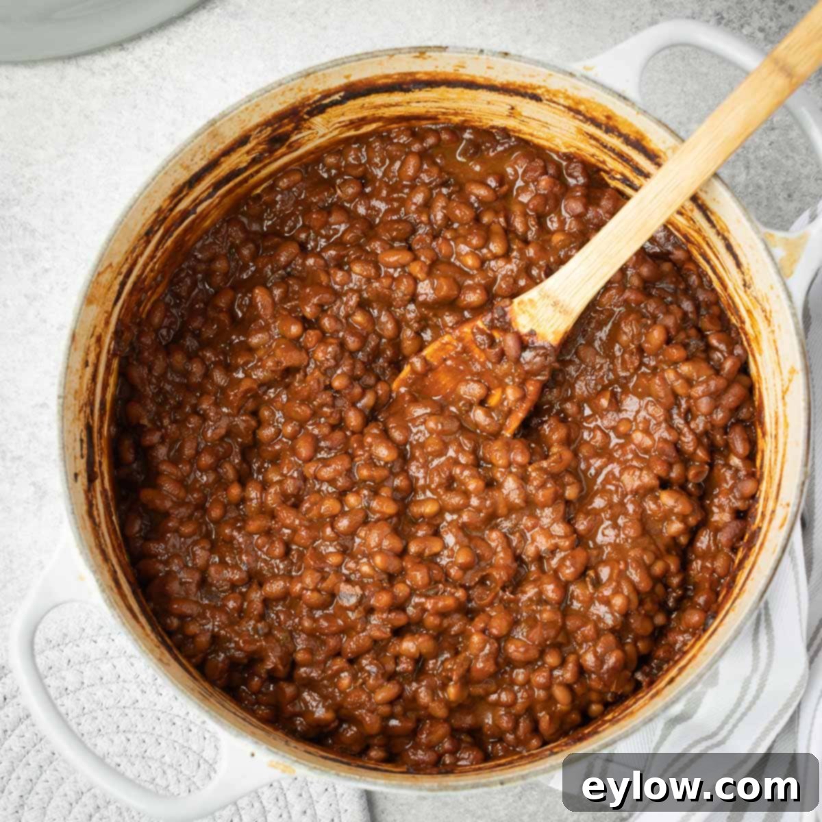 A gray Dutch oven full of savory-sweet dark brown baked beans, with a wooden spoon resting inside, showcasing the rich texture.