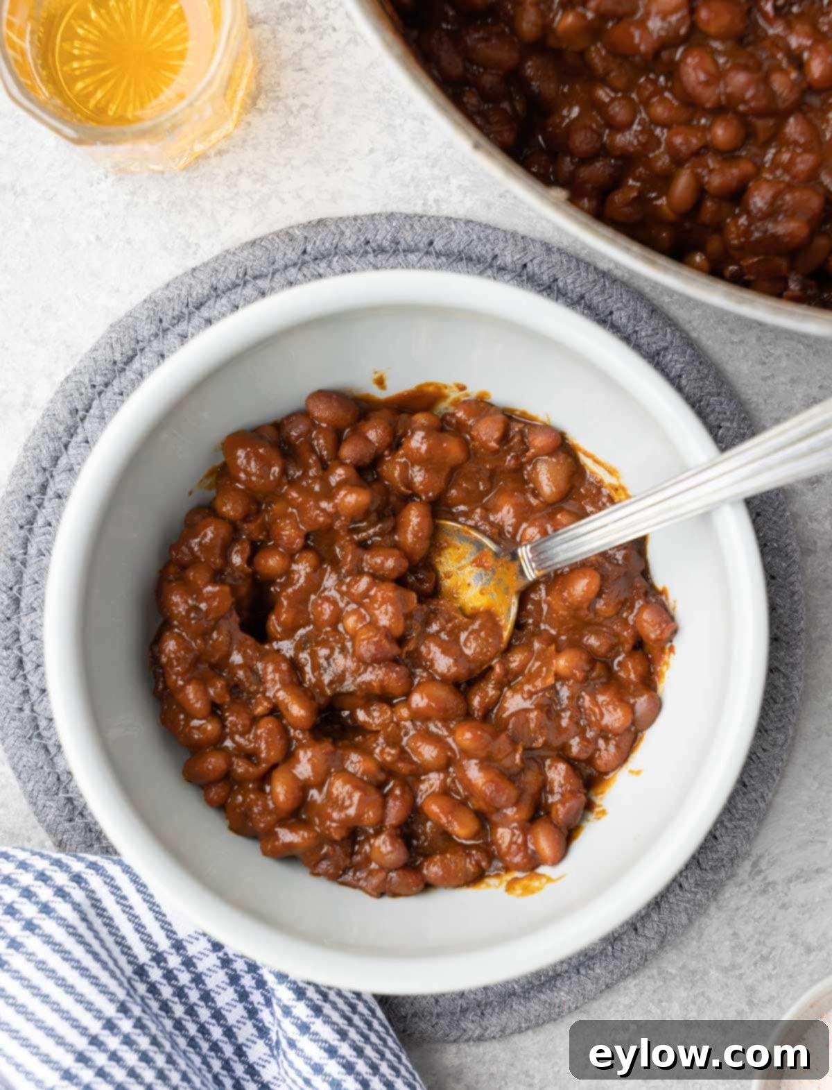 A small bowl of deep brown Boston baked beans, garnished lightly, with a blue checked towel and wooden spoon nearby, suggesting a comforting meal.