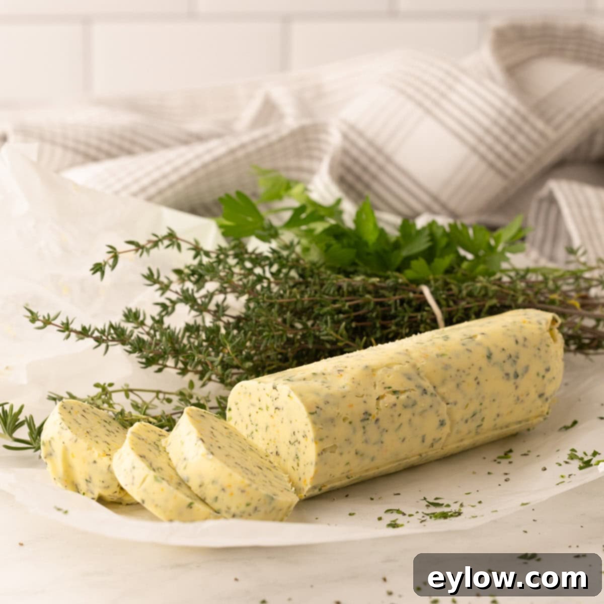 A yellow butter log with herbs with sliced pieces, fresh herbs behind.