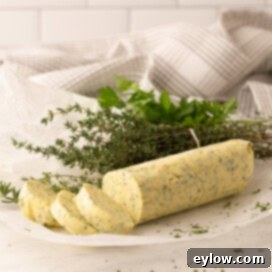 A yellow butter log with herbs with sliced pieces, fresh herbs behind.