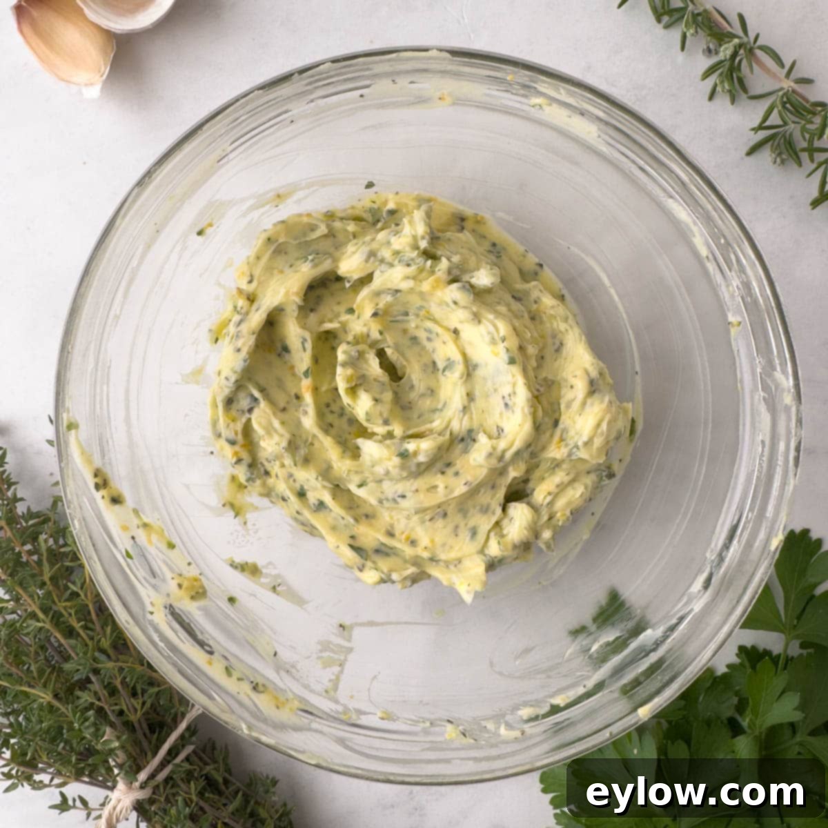 A soft compound butter mixed with herbs in a glass bowl with herbs on side.