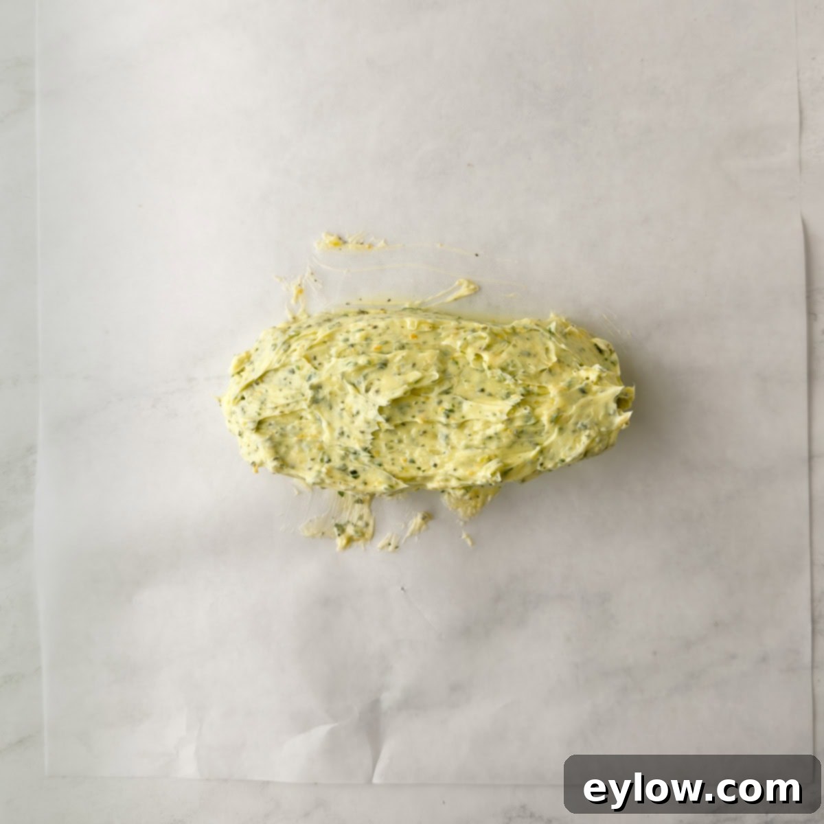 A yellow compound butter with flecks of green herbs on a piece of white parchment paper. 