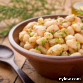 A simple white bean salad in a wooden bowl with bits of lemon zest, herbs, and carrots and a serving spoon near.