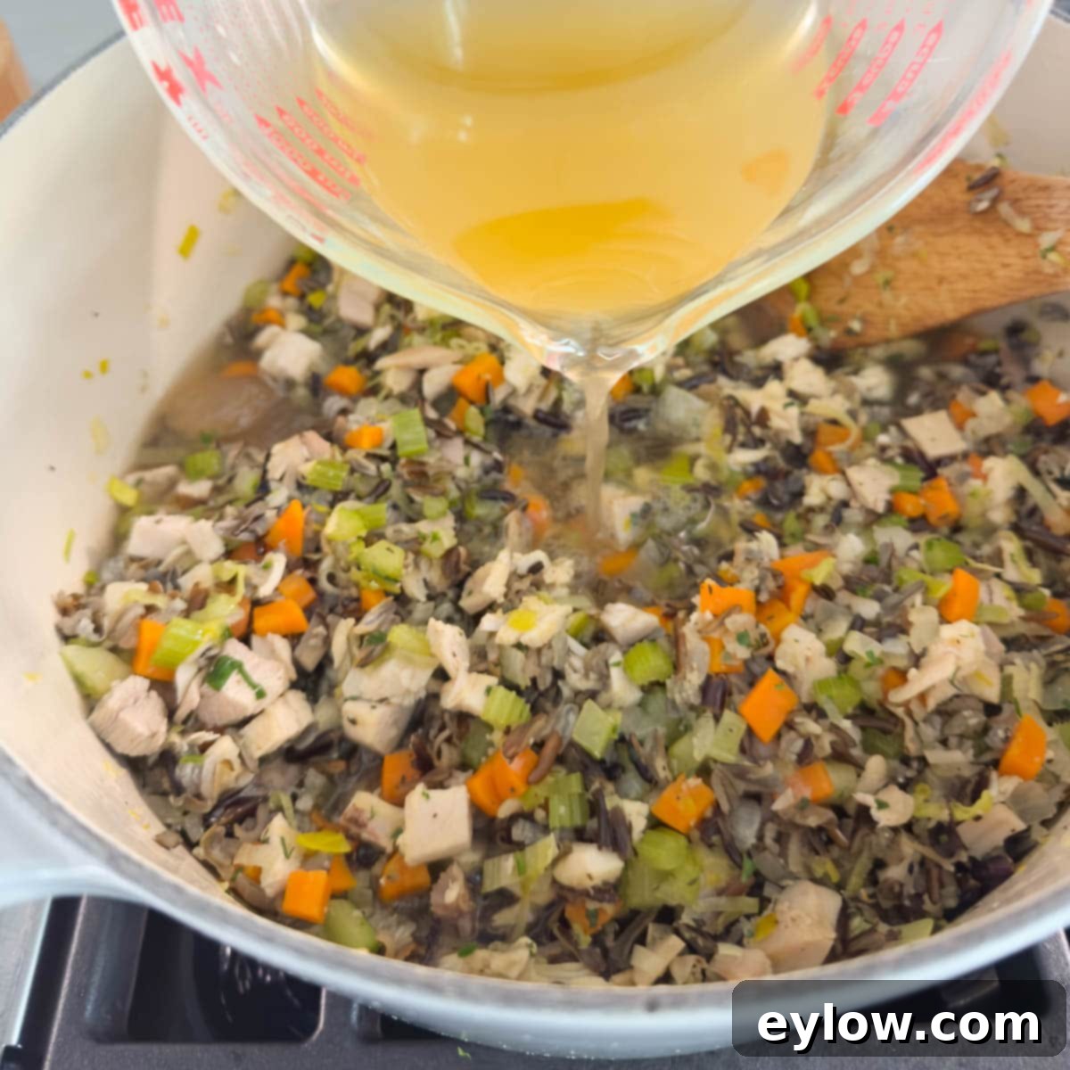 Pouring rich golden broth from a large glass measuring cup into a pot of simmering wild rice soup with turkey and vegetables.