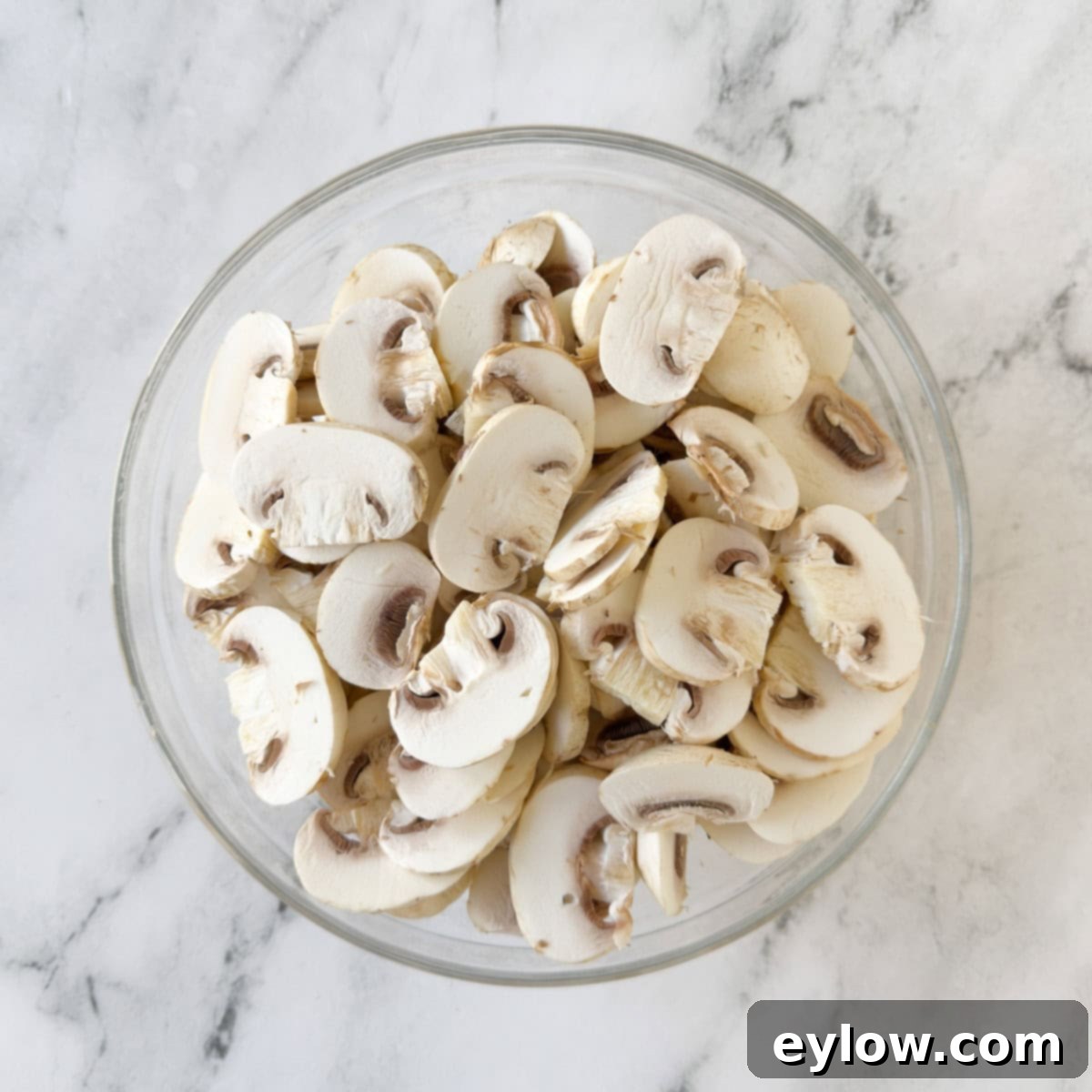 A glass bowl of sliced white mushrooms on a marble counter.