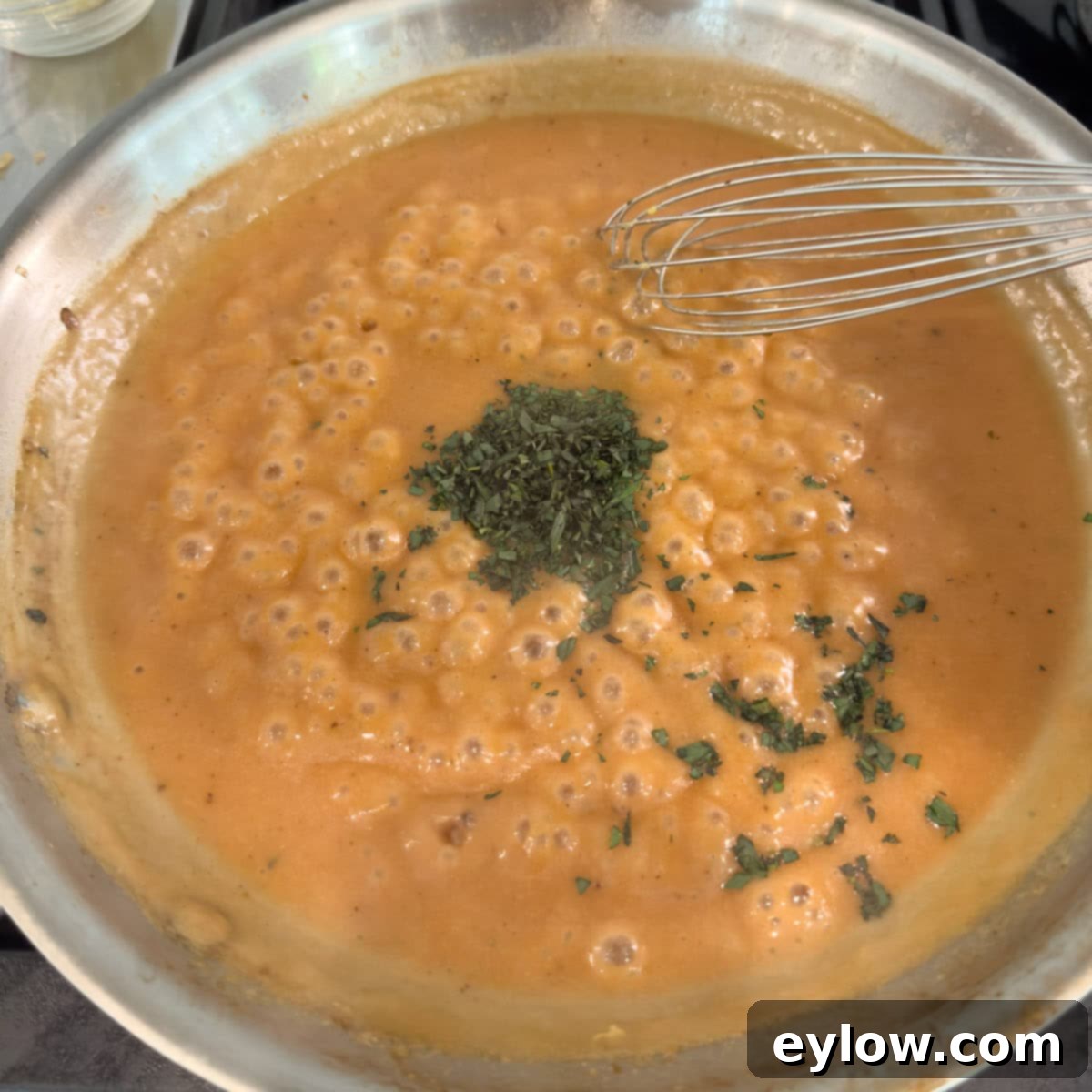 A bubbling pan of stroganoff sauce in the making, adding herbs with a whisk.