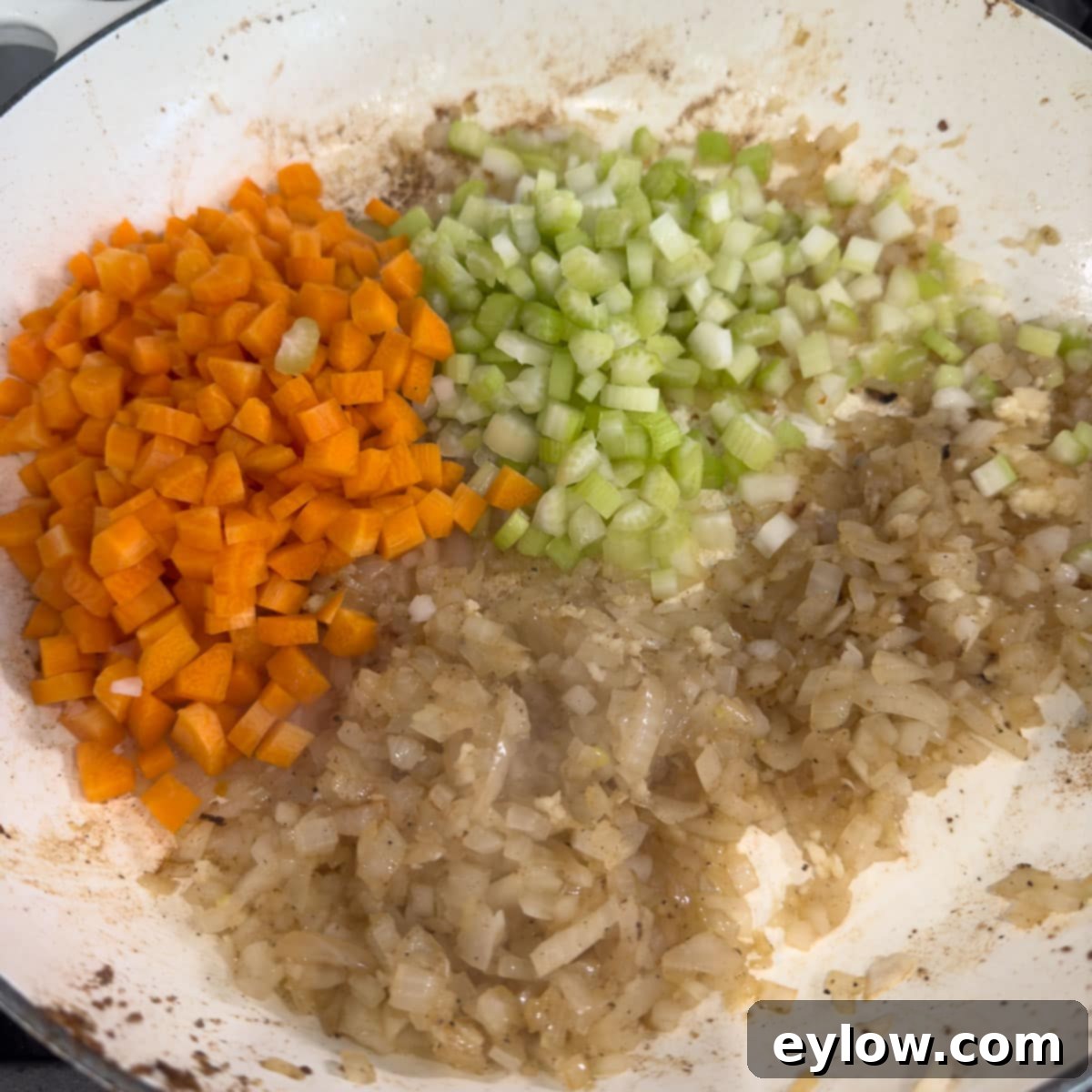 Cooking chopped onion, carrots, and celery in a white wide shallow pan.