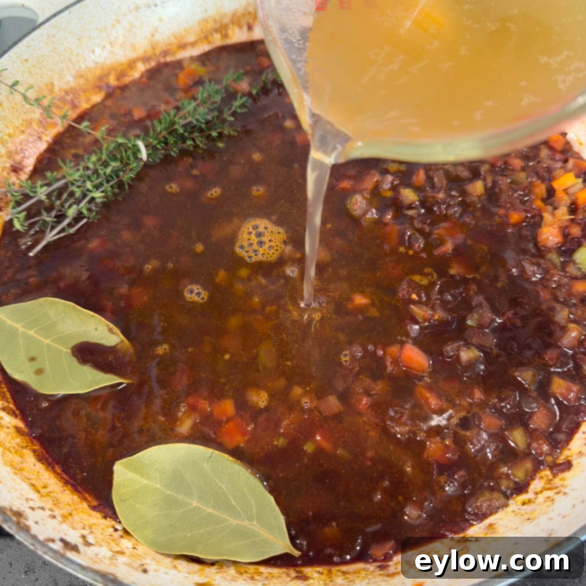 pouring broth into a pan of vegetables and wine with bay leaves and thyme.
