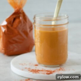 A glass jar of bright orange paprika sauce with a golden spoon and a bag of paprika behind.