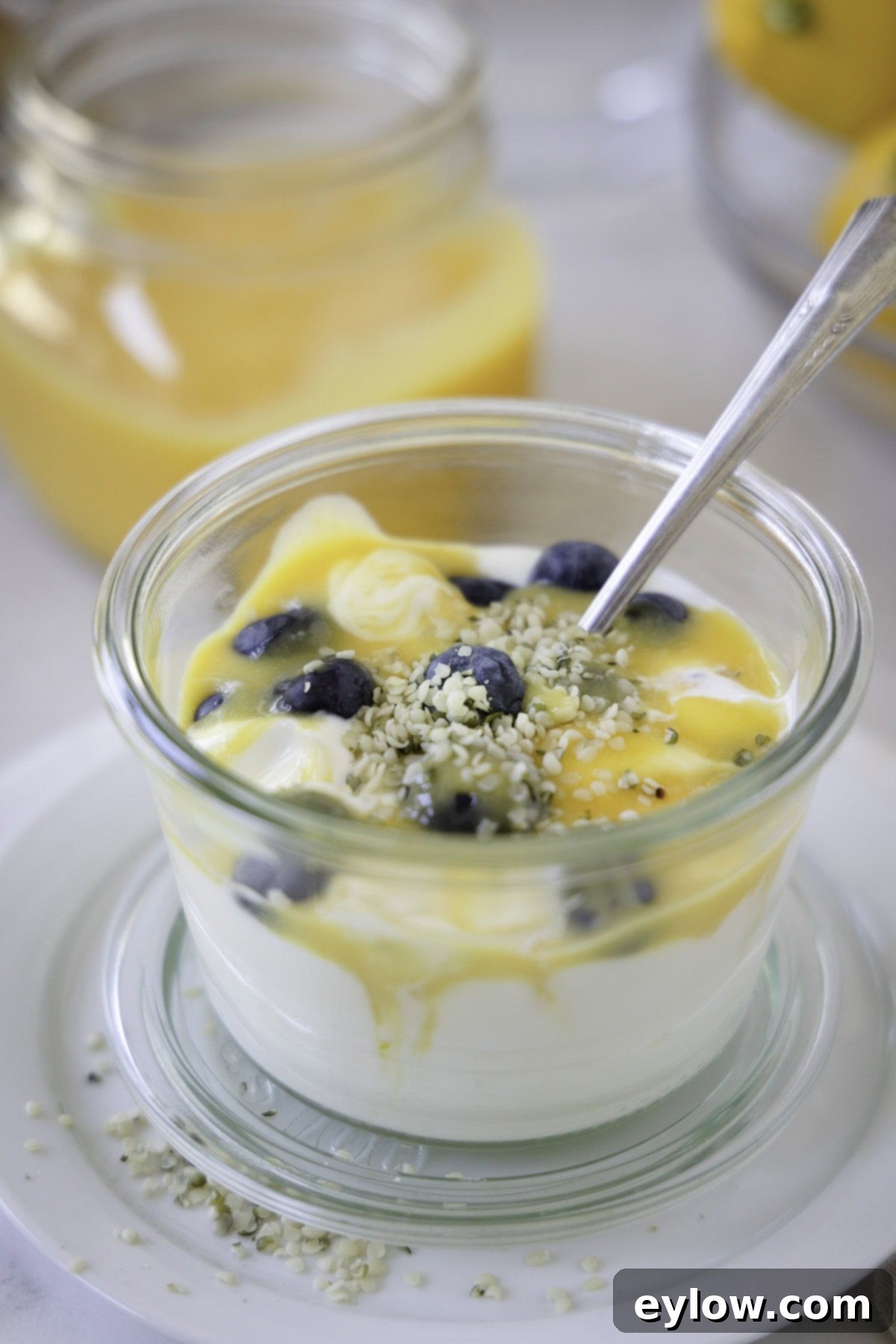 A jar of creamy yogurt topped generously with bright yellow sugar-free lemon curd and fresh blueberries, with a silver spoon ready to dig in.