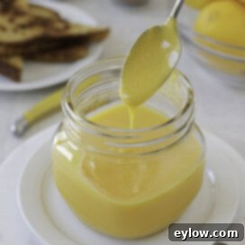 A square glass jar brimming with luscious lemon curd, with a silver spoon capturing a perfect drip, emphasizing its creamy texture.