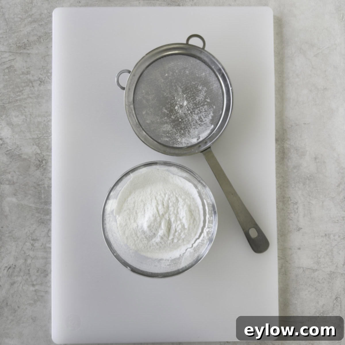 A stainless steel small sieve with finely sifted powdered allulose on a white cutting board, ready for use.