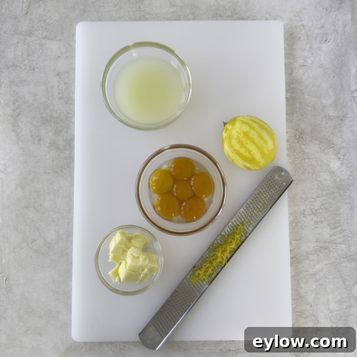 Various prepped ingredients for sugar-free lemon curd arranged on a cutting board: fresh lemon juice, cubed unsalted butter, and vibrant egg yolks.