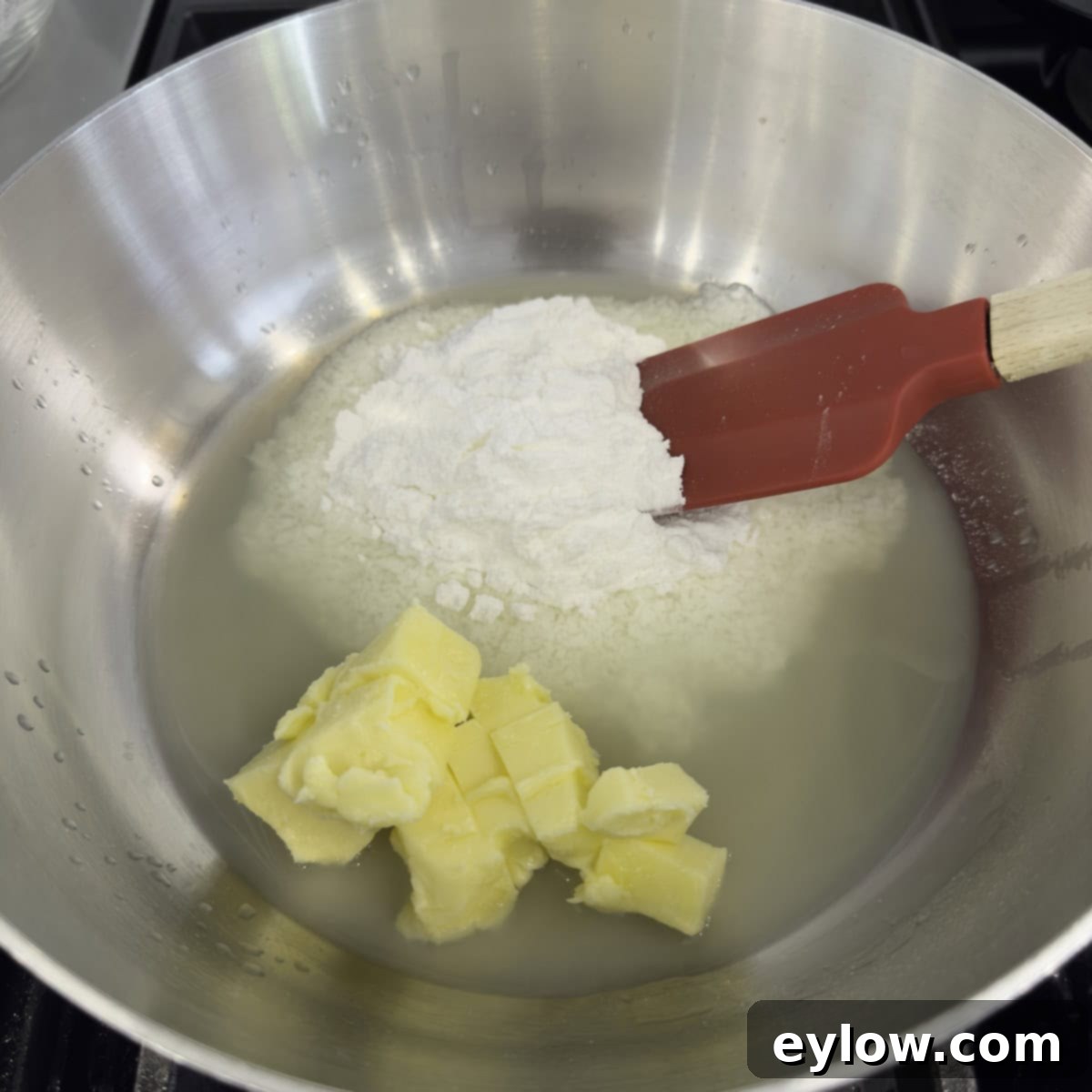 A stainless steel saucepan on a stovetop, containing lemon juice, cubed unsalted butter, and sifted allulose, heating gently to melt.