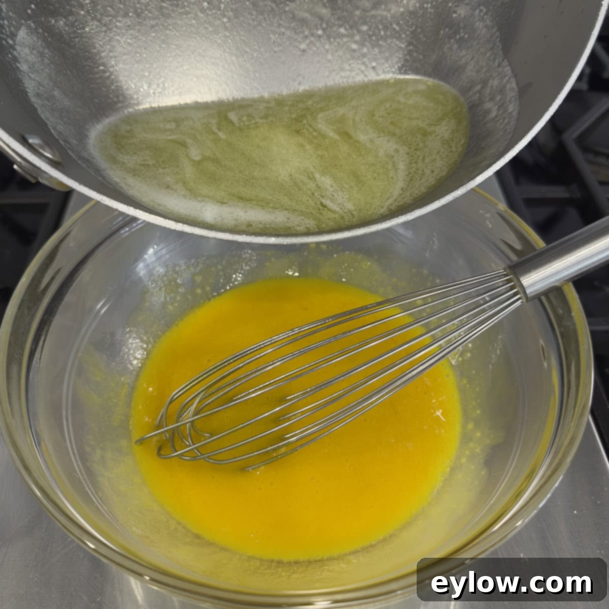 A stream of hot lemon-butter-allulose mixture being slowly drizzled into a bowl of bright yellow egg yolks, with a whisk in action to temper the eggs.