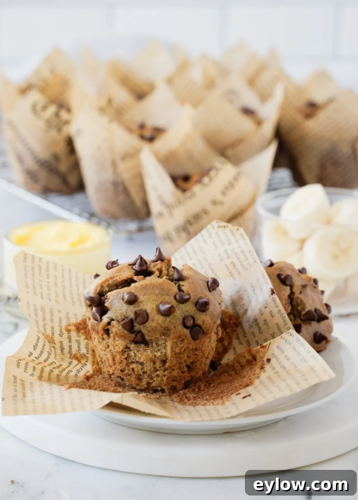 Delicious gluten-free chocolate chip banana muffins presented on a small plate, with more muffins in elegant tulip paper wraps arranged in the background. The muffins are golden brown with visible chocolate chips, promising a tender and moist bite.