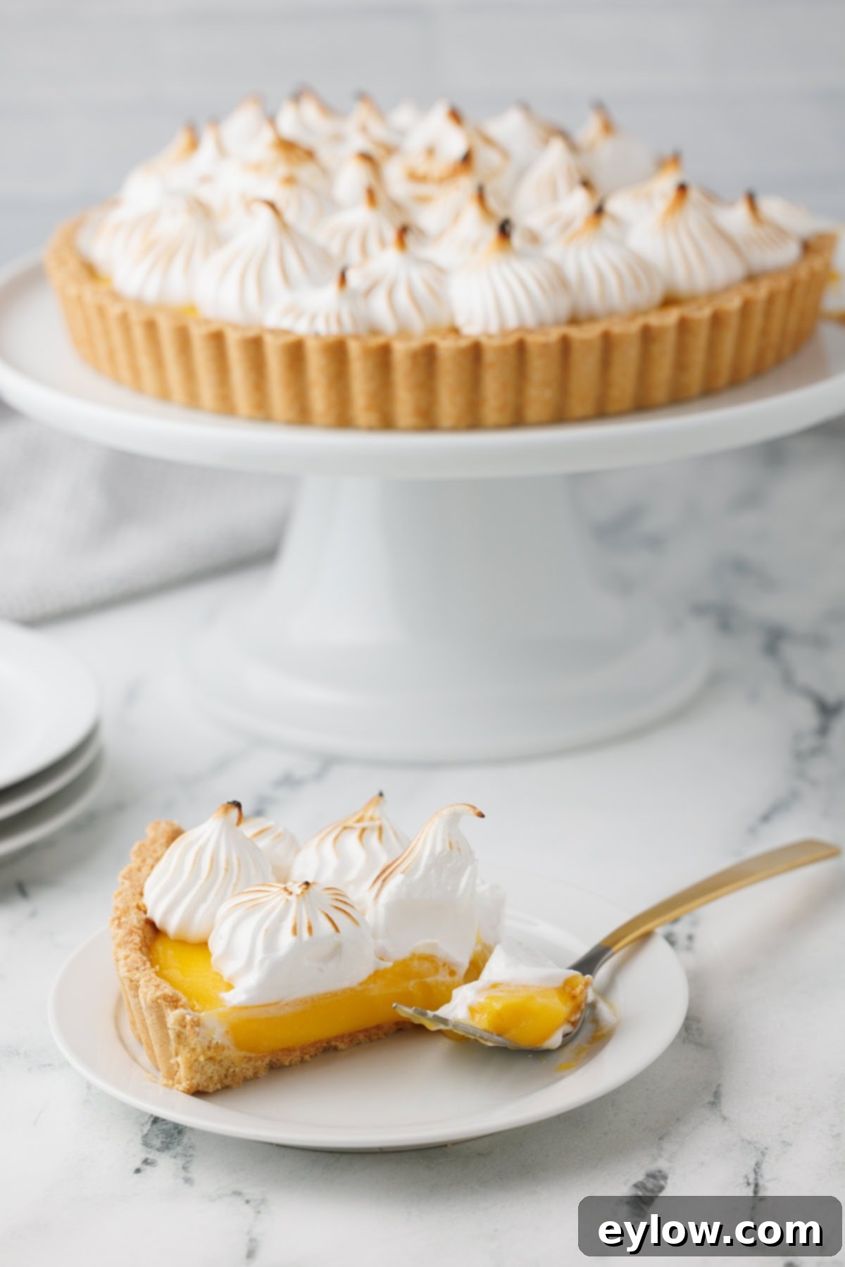 Zesty Lemon Meringue Tart 13 A lemon meringue tart on a white cake stand with a slice of tart on a small plate in front, garnished with a mint sprig.